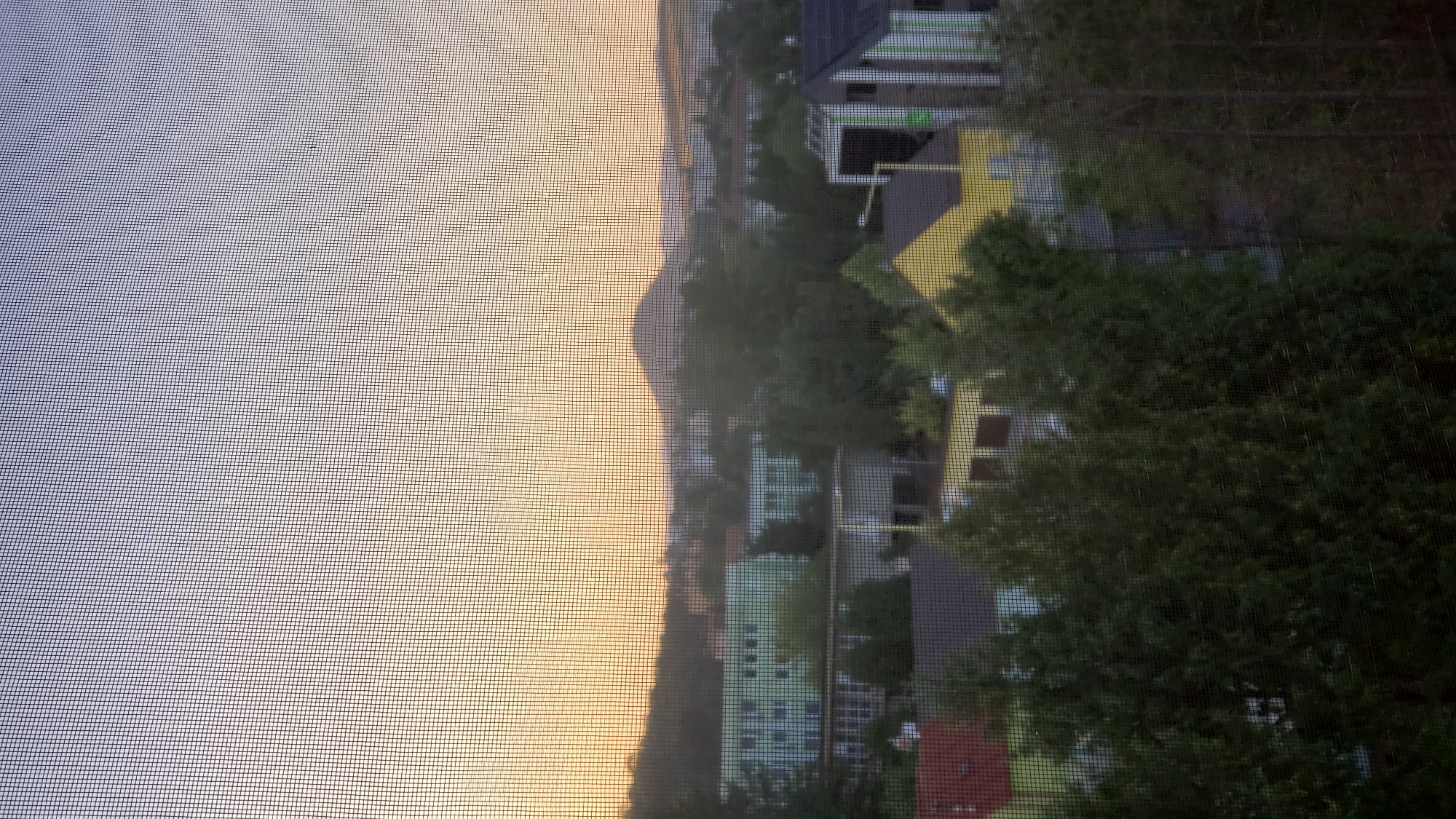 The image depicts a view of Prešov, Slovakia, through a window screen at dusk. The sky is a gradient of soft blues and oranges, suggesting the sun has recently set or is about to rise. In the distance, a prominent conical hill, likely the Zlatá Baňa volcano, rises above the cityscape. Numerous buildings are visible, including apartment complexes with green facades, smaller residential houses, and what appears to be a school or public building with a yellow roof. Lush green trees are interspersed throughout the scene, obscuring some of the structures and contributing to a suburban or semi-rural feel within the city. The overall impression is a quiet, residential area viewed from an elevated position.
