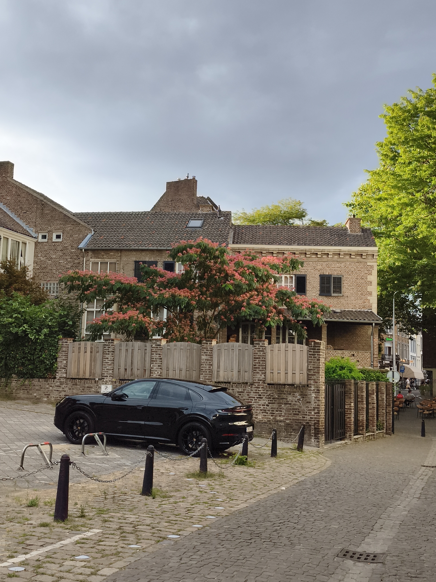 The image depicts a street scene in the city of Maastricht, Netherlands. In the foreground, a black car is parked on a cobblestone surface, partially obscured by metal parking barriers and chains. Adjacent to the parking area is a brick wall with a wooden fence. Behind the fence, a building with brickwork and tiled roofs is visible, featuring windows and shutters. A tree with pink blossoms is situated in front of the building. To the right of the building, a street continues, lined with tables and chairs outside what appears to be a cafe or restaurant. In the background, other buildings and trees are visible under a cloudy sky.