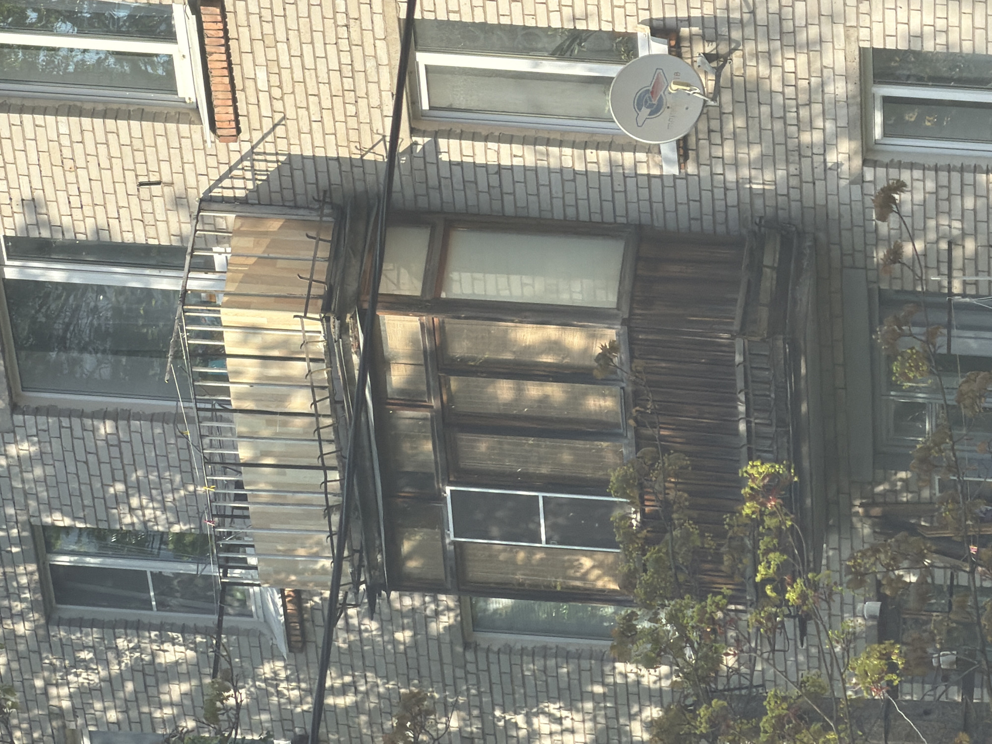 The image shows the exterior of a brick building with several windows. A wooden balcony or enclosed porch with windows is visible on the building's facade. A satellite dish is mounted on the wall above the balcony. A metal fire escape ladder runs alongside the building. The exact location in Tokkari-Lend, Russia, is not identifiable from the image.