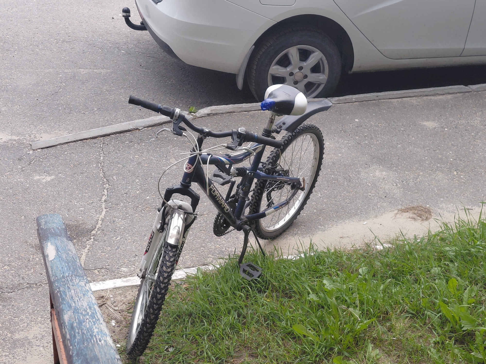 In Nefteyugansk, Russia, a dark blue bicycle with a black seat and white accents is parked on a paved surface next to a patch of green grass. The bicycle has silver-colored handlebars, front fork, and fenders. A blue plastic bag is tied around the seat post. The bicycle is leaning on its kickstand, which is positioned in the grass. Behind the bicycle, a white car is partially visible, with its rear wheel and lower body showing. The car has a trailer hitch attached to its bumper. The paved area appears to be a street or parking lot, with visible cracks in the asphalt. A weathered blue and brown wooden object, possibly a bench or a plank, is located to the left of the bicycle. The scene is outdoors during the daytime, under an overcast sky.