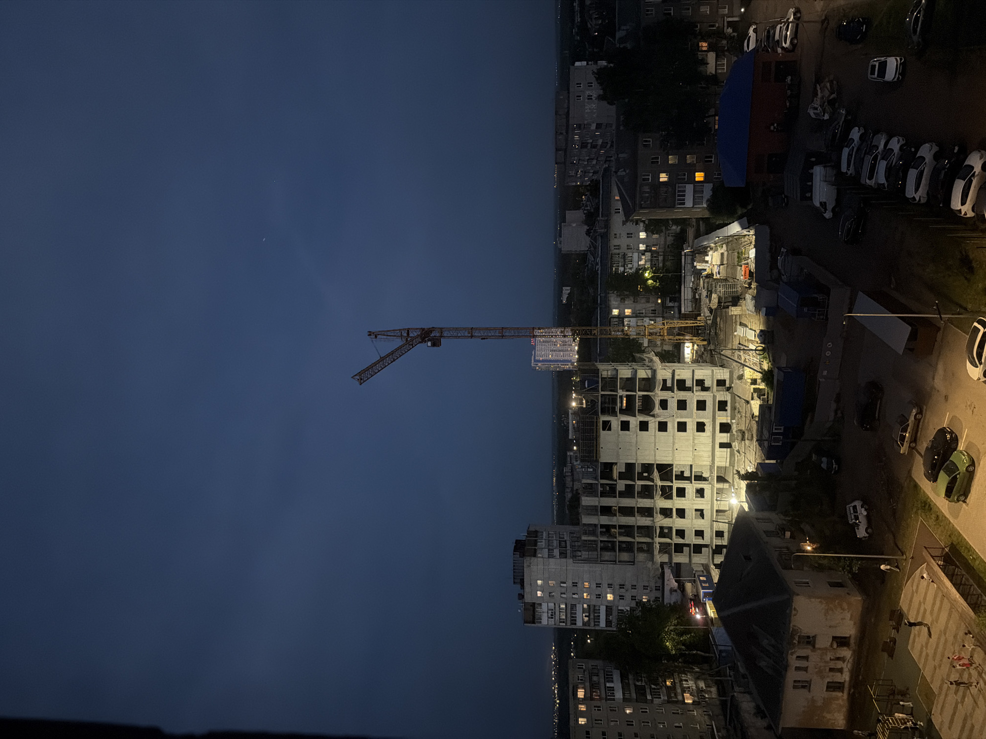 The image shows an evening cityscape. A tall construction crane stands near an unfinished high-rise building. Surrounding buildings are lit with artificial light. To the right of the construction site is a parking area with parked cars. People are visible in a lighted square on the bottom right. The sky is dark blue, indicating nighttime.