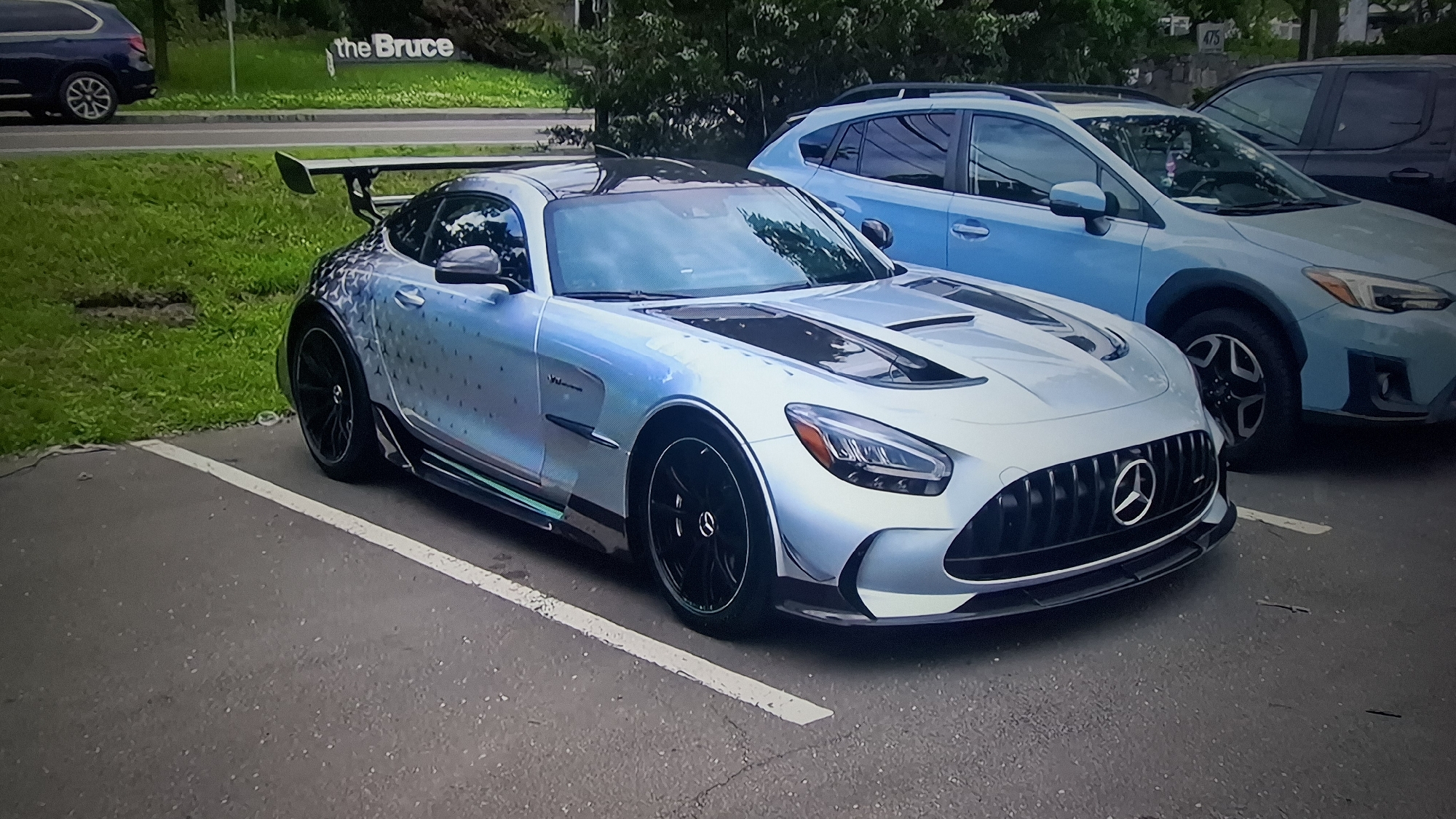 A light blue or silver Mercedes-AMG GT R, featuring a distinctive patterned wrap composed of numerous small star-like shapes on its rear half and sides, is prominently parked in an asphalt lot. The car has black multi-spoke wheels, a large black rear spoiler, and black accenting on its hood. A green and black striped detail runs along its side sills. Badges such as "AMG" and "V8 BiTurbo" are visible on its side.
To the right of the Mercedes, a light blue Subaru Crosstrek or similar SUV is parked. Partially visible behind the Subaru is a dark-colored SUV or truck. On the far left of the image, the rear section of a dark blue SUV is visible, suggesting a row of parked vehicles.
The parking lot features white painted lines. Beyond the cars, there is a green grassy verge leading to a road. Trees and bushes form a dense green background. A white sign with the text "the Bruce" is visible amidst the foliage behind the road. Faintly, the numbers "475" can be seen on a building or sign in the background near the Subaru. The scene takes place during the daytime, under what appears to be overcast or shaded conditions, as no strong shadows are present. No people are visible in the image.