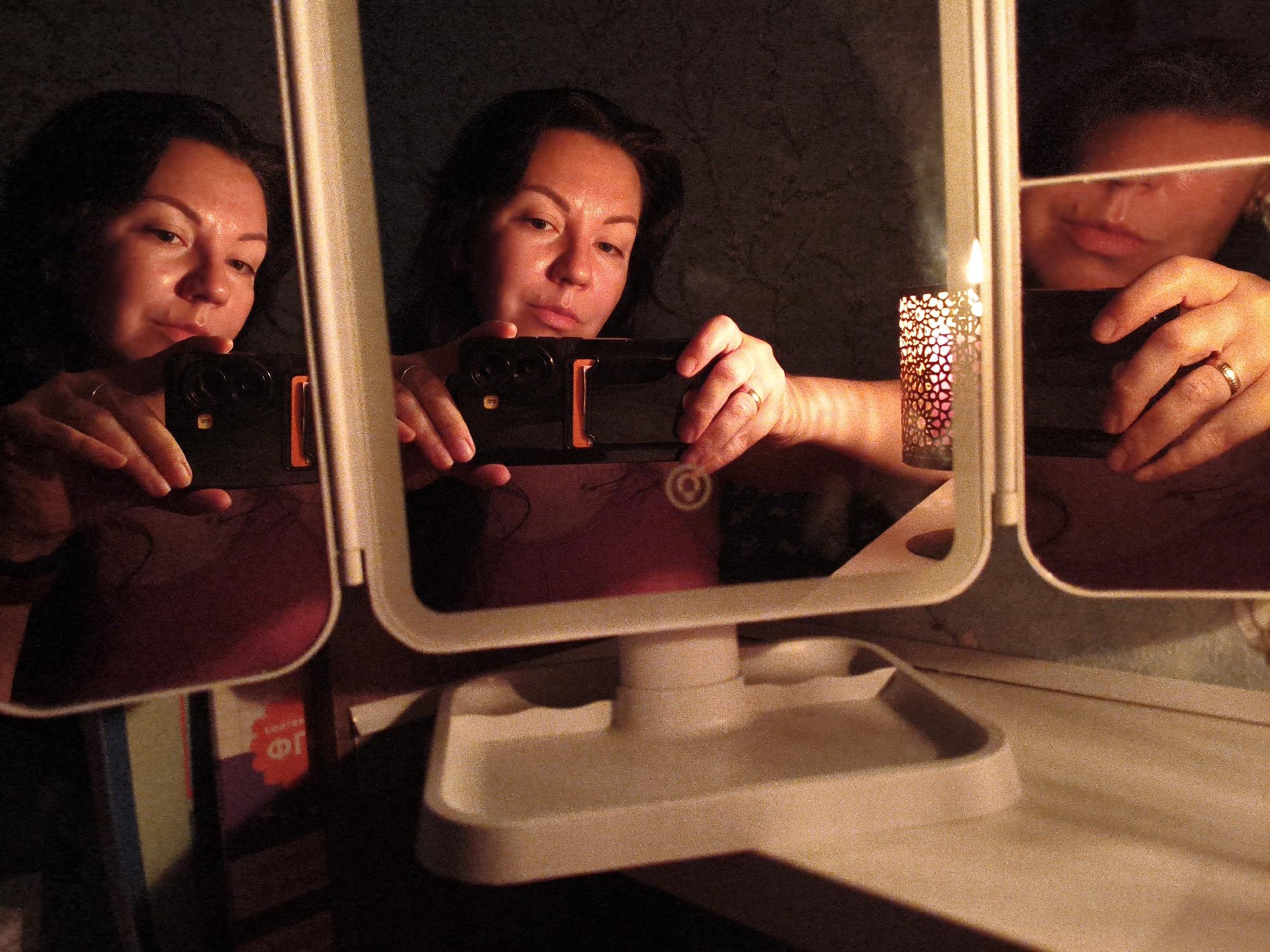 A woman is taking a selfie in a mirror, holding a smartphone in her hands. A lit candle in a decorative holder is visible to the right of the mirror. The scene appears to be indoors, possibly a bathroom or bedroom, given the presence of a mirror and what looks like a vanity or shelf. The lighting is dim, with the candle providing a warm glow. The woman is wearing jewelry, including rings on her fingers. The overall atmosphere is intimate and personal, suggesting a quiet moment captured by the woman. The city of Orenburg, Russia, is mentioned as the location, but there are no specific visual cues in the image to confirm this.