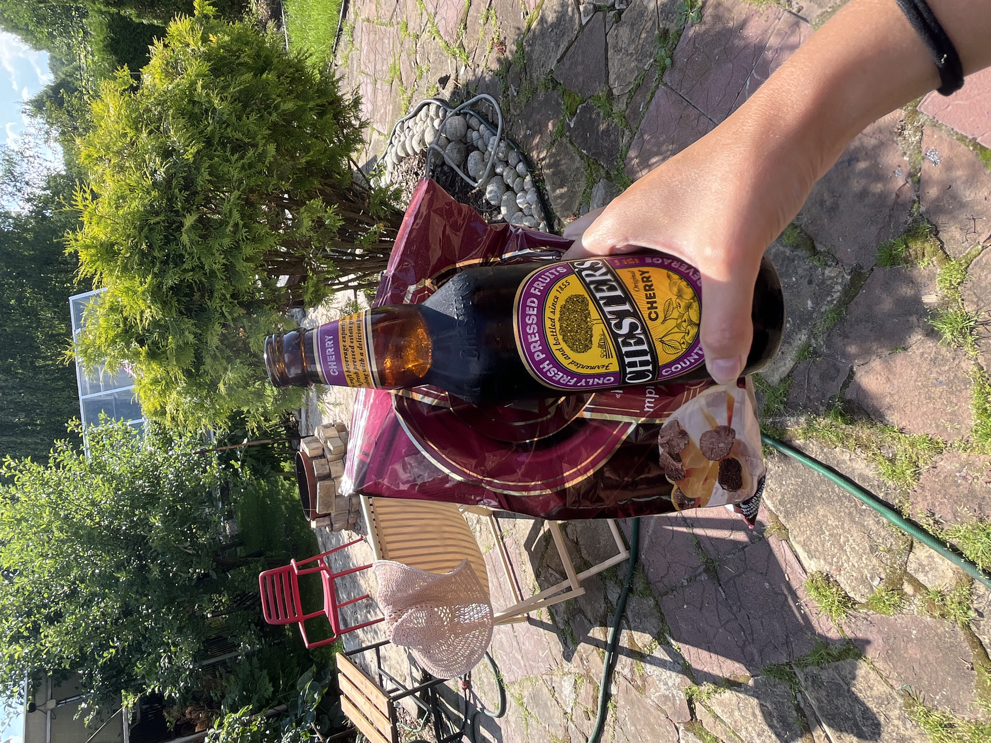 In the city of Вельяминово, Russia, a person's hand holds a bottle of Chesters Cherry beverage and a partially open bag of chocolates outdoors. The scene is set in a backyard with a paved patio, scattered stones, and green grass. Sunlight casts shadows from trees and other objects. In the background, there is a red chair, a wooden table with chairs, and a greenhouse. The overall impression is one of a casual, sunny day.