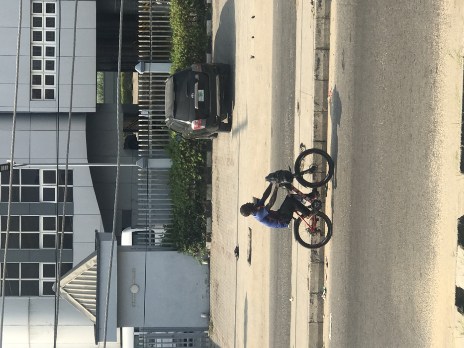 An outdoor scene in an urban area, identified as Ado, Nigeria, depicts a male individual on a red and black mountain bicycle positioned on a paved shoulder adjacent to an asphalt road. The cyclist, wearing a blue and red striped short-sleeved shirt (resembling an FC Barcelona jersey), dark trousers, and dark footwear, faces slightly right, appearing to be in motion or paused. A black bag is attached to the bicycle's handlebars. Litter, including small paper fragments and dark debris, is visible on the ground and curb near the bicycle.
In the background, a light grey concrete gatehouse with a triangular metal roof stands on the left. Behind this structure and a grey metal fence, a white van is partially visible. To the right of the gatehouse, behind the fence and a row of green bushes, a black Toyota Highlander SUV is parked, facing away from the viewer, on a paved lot. Dominating the upper background is a multi-story modern building with a grey and silver facade, featuring numerous square windows and multiple horizontal power lines traversing the upper frame. The scene is illuminated by strong daylight, casting distinct shadows from the bicycle and the parked SUV.