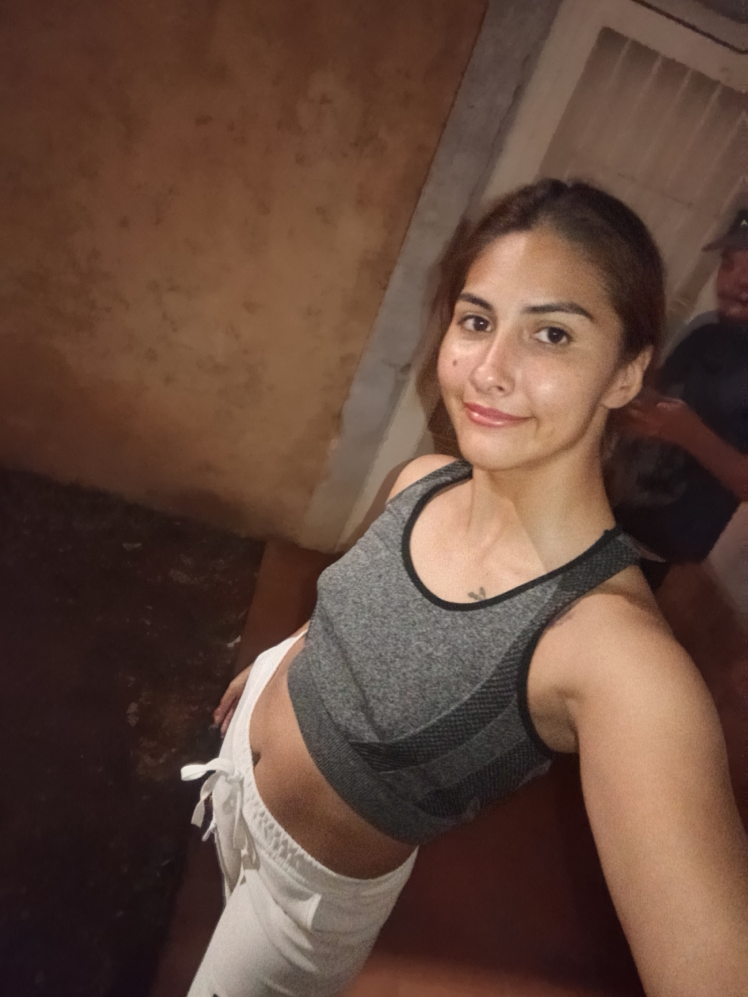 This is an indoor, eye-level selfie featuring a young woman in the foreground, with a man partially visible in the background. The woman, who has medium brown hair pulled back, is smiling gently at the camera. She has a fair complexion with visible freckles or blemishes and light-colored lips. She is wearing a grey sports bra with black trim and white, loose-fitting pants tied at the waist, exposing her midriff. A small, dark tattoo is visible on her upper left chest.
Behind her, to the right, a man's upper body is partially visible. He is wearing a dark shirt and a dark baseball cap, with his face mostly obscured. The setting appears to be a simple interior space, possibly a home or hallway in Juan Griego, Venezuela. The wall on the left is a textured, mottled reddish-brown color, while the floor is a dark, possibly concrete, surface. There's an indistinct corner or doorway to the right. The lighting is warm and slightly dim, suggesting artificial illumination, possibly evening or nighttime. The overall atmosphere is casual and candid. There is no visible text in the image.