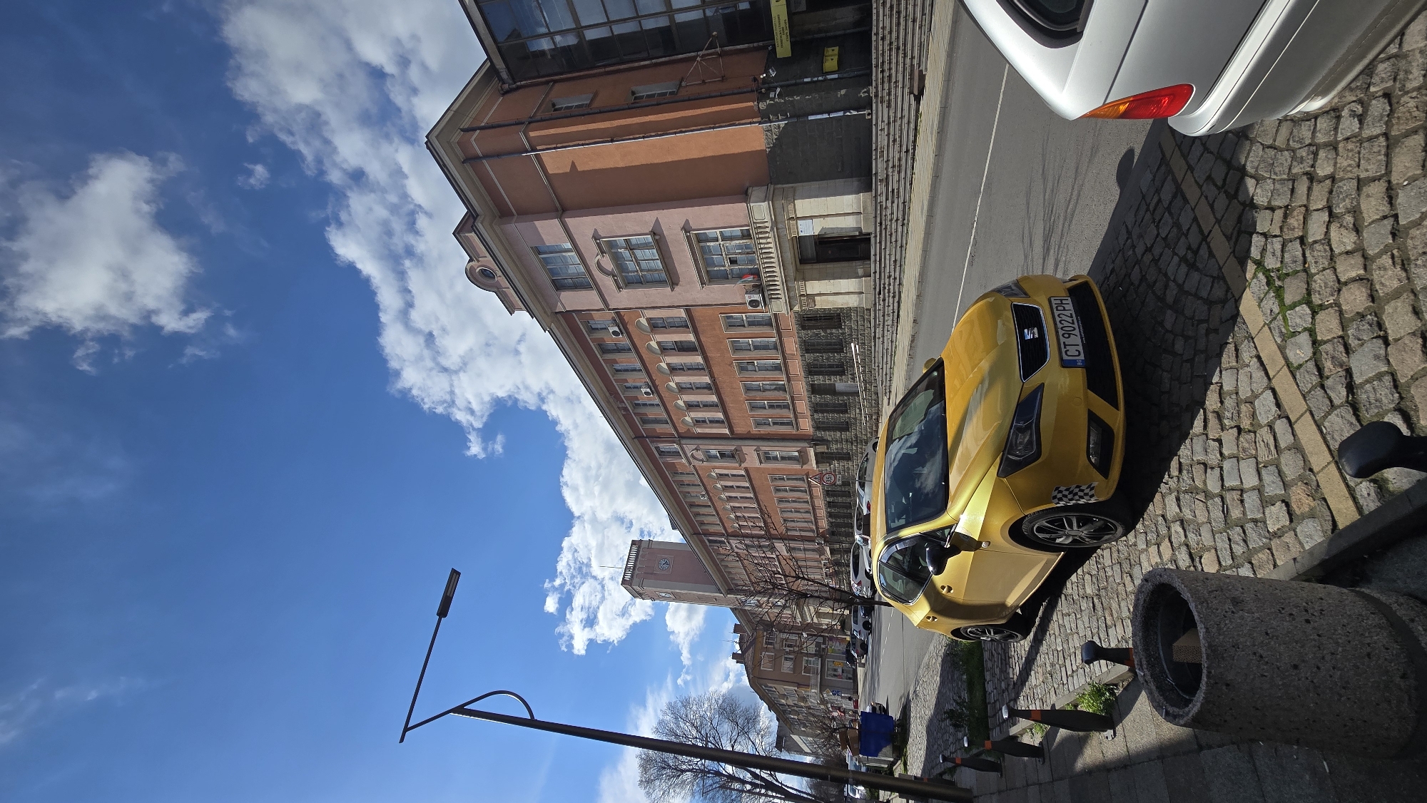 The image captures a street scene in Stara Zagora, Bulgaria. A yellow Seat car is parked on a cobblestone street adjacent to a sidewalk. Next to it is a concrete block which serves as a barrier, with additional traffic cones. Parked nearby is a white car. The buildings along the street are a mix of historical and modern architecture, predominantly several stories in height. The sky is blue with scattered white clouds. A street lamp is visible in the foreground. Bare trees are also present. The building that seems to be near the center is the Regional Directorate of Forests Stara Zagora.