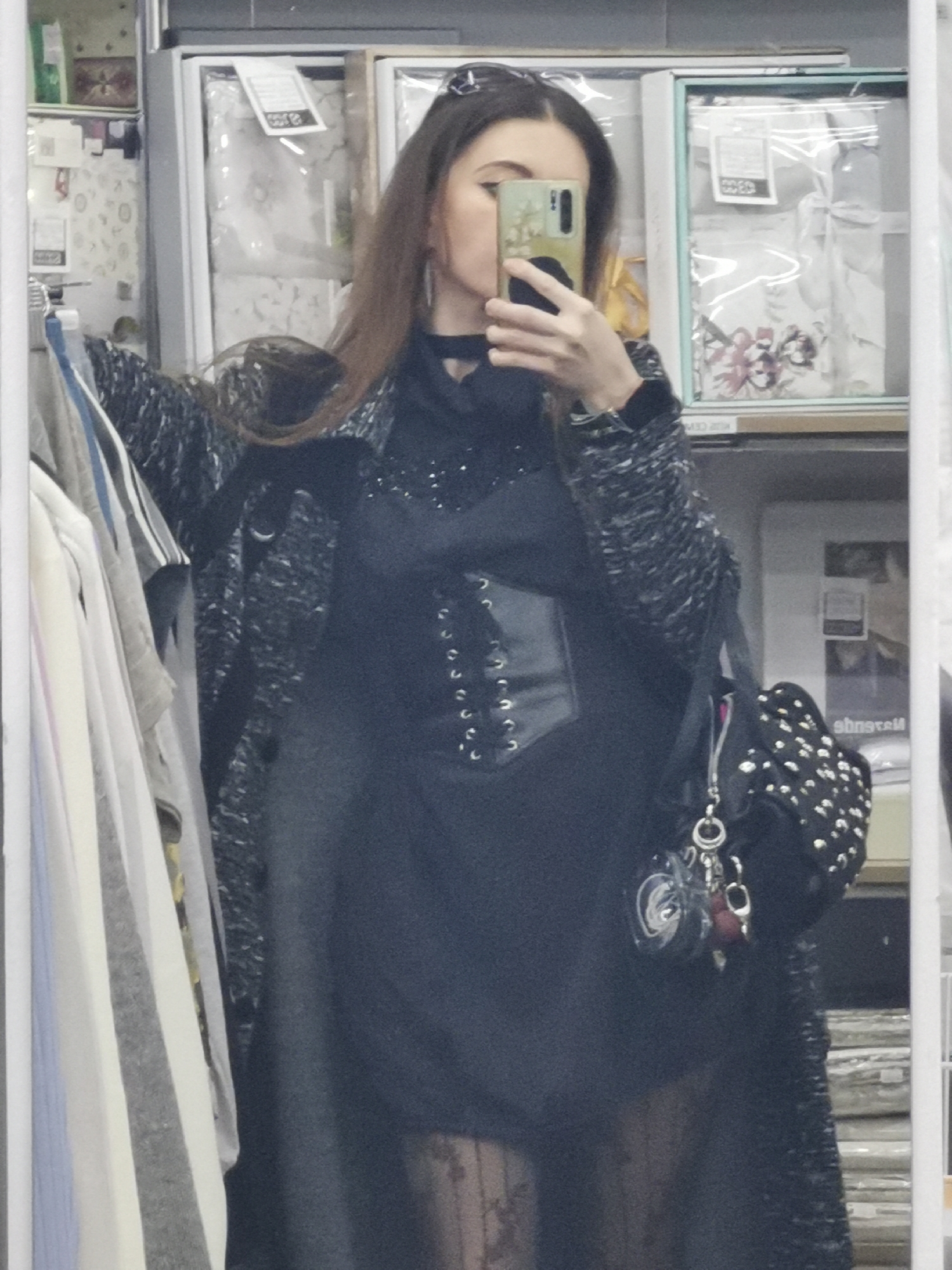 A person stands in front of a mirror, taking a selfie with a smartphone. The person is wearing a black outfit with a corset-style bodice, a textured coat, and a handbag with studded detailing. The reflection suggests the person is in a retail store, with merchandise like packaged bedding visible behind them. The location is in an unspecified urban setting, as indicated by the retail environment and the presence of goods for sale.