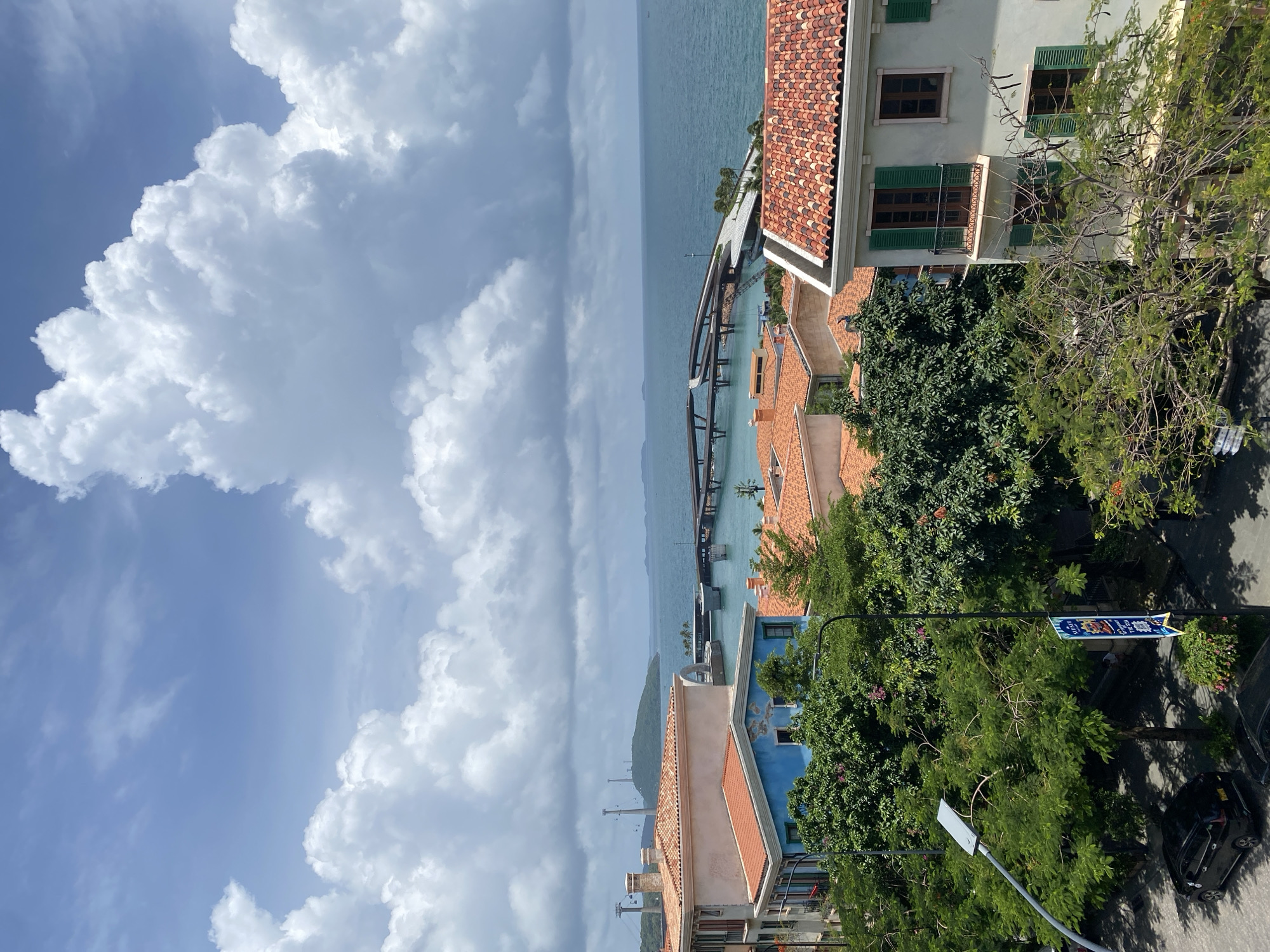 This image captures a view of the coastal city of Phú Quốc, Vietnam, featuring a blend of European-inspired architecture and the natural beauty of the ocean and sky. In the foreground, several buildings with terracotta tile roofs and pastel-colored facades are visible, characteristic of Mediterranean design. Lush green trees with vibrant foliage dot the landscape, interspersed with the buildings and a street scene where a black car is parked. A modern streetlamp with a prominent light fixture stands tall among the trees.
Beyond the buildings, the azure waters of the ocean stretch to the horizon under a bright, partially cloudy sky. A distinctive bridge structure, with an elevated and curving design, spans across the water, connecting different parts of the coastline. In the distance, a verdant island or landmass can be seen, with a faint indication of infrastructure like towers and cable cars, suggesting a developed resort or entertainment area. The overall scene evokes a sense of a vacation destination, where architectural charm meets coastal serenity.