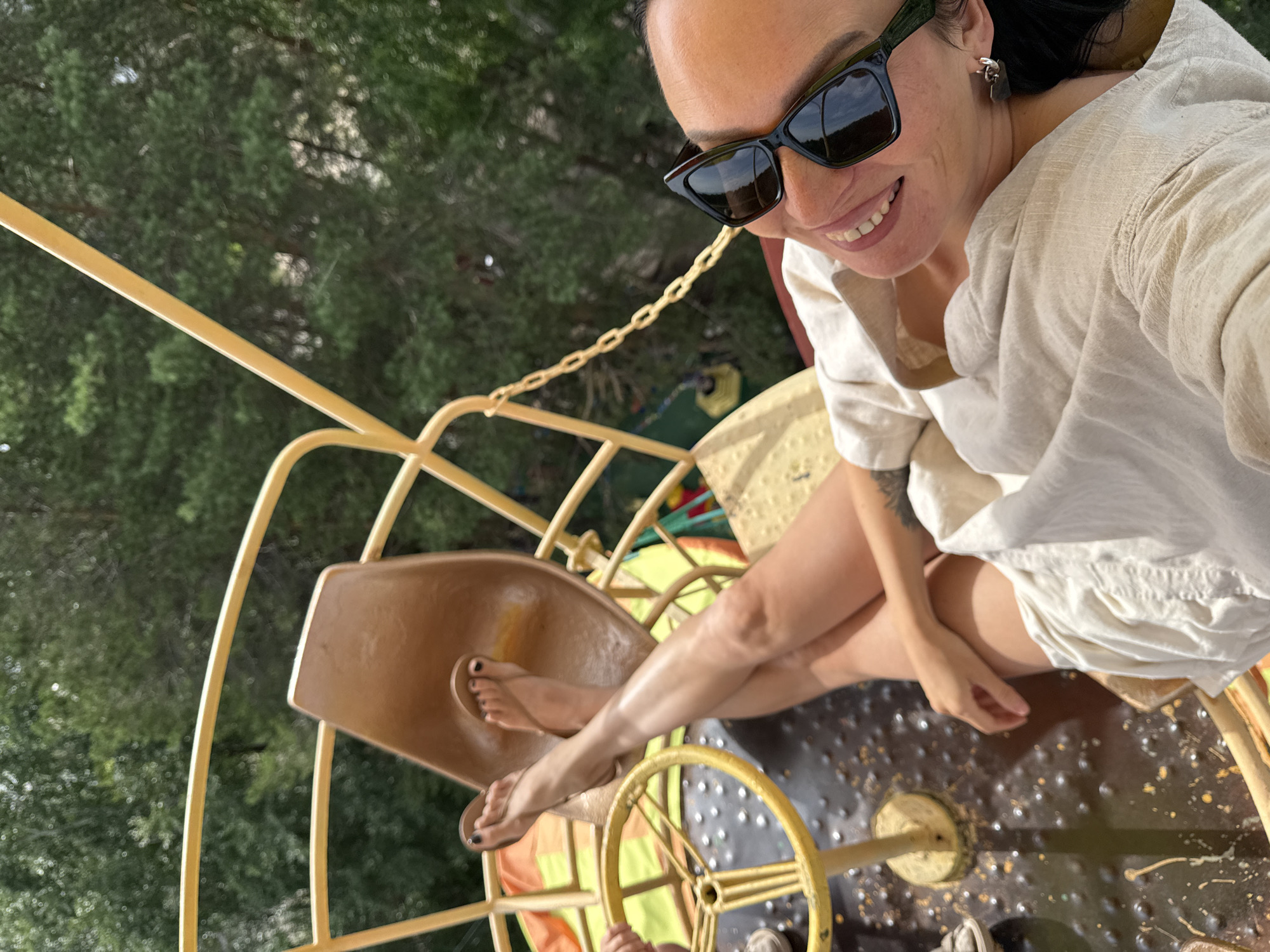 A high-angle, medium shot shows a woman sitting in a yellow amusement ride, possibly a carousel or a similar attraction, with a blurred background of green trees. The woman is wearing sunglasses, a light-colored, loose-fitting outfit, and appears to be smiling. Her legs are crossed, and one foot, adorned with black nail polish, is resting on a part of the ride. The ride itself has a metallic yellow frame and a brown seat. The ground beneath the ride is dark and speckled. The lighting suggests it is daytime, and the foliage indicates an outdoor setting, likely in a park or recreational area. The overall mood is cheerful and relaxed. There is no visible text in the image.
