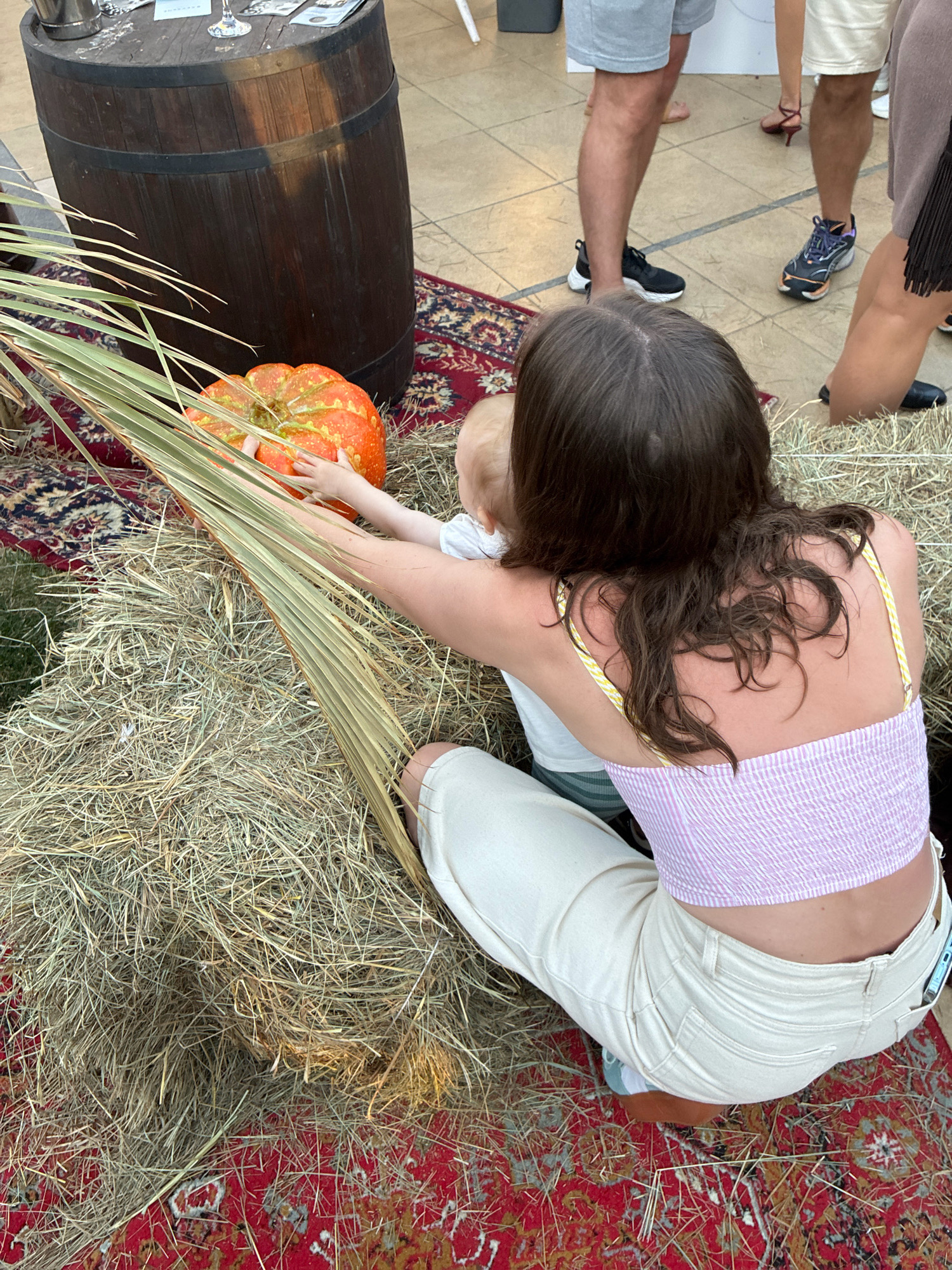 In Sochi, Russia, on a patterned rug laid over a paved surface, a woman is crouched next to a bale of hay. She wears a light-colored crop top with a pink striped pattern and matching light-colored pants. Her back is to the camera, and she is reaching towards a small, orange pumpkin with a mottled green and yellow pattern. Beside the pumpkin, a baby wearing a white shirt is reaching for it as well. A dried palm frond extends diagonally across the scene from the left. In the background, a dark wooden barrel with metal bands stands on the rug, and several adults are visible, some wearing casual attire. The overall impression is a festive or autumnal gathering.