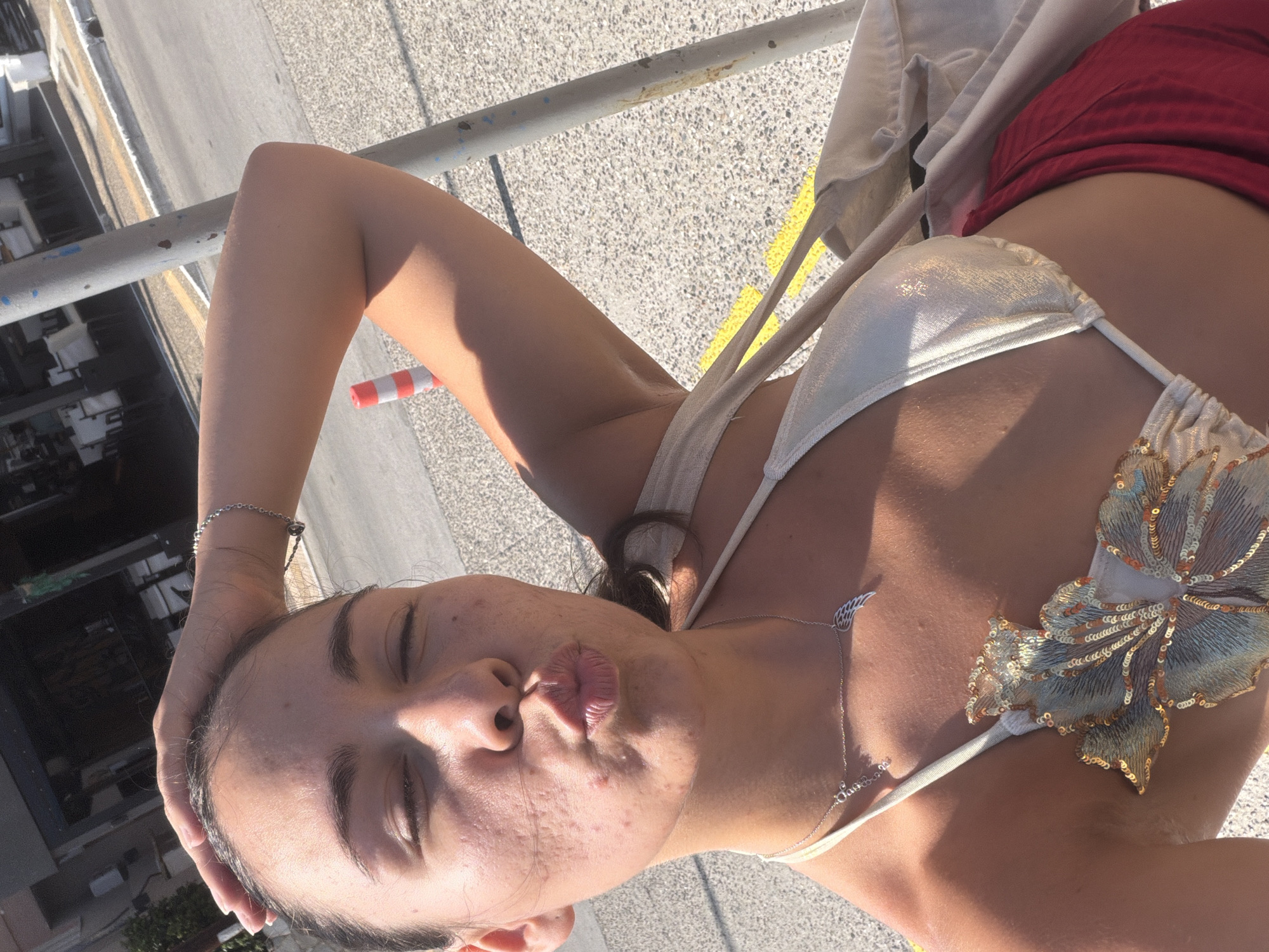 A young woman is pictured in a close-up, tilted shot, outdoors on a sunny day. She has dark hair and is wearing a white bikini top with a gold floral applique and a red skirt or shorts. A silver necklace with a wing charm and a small pendant is visible. Her left arm is raised, with her hand resting on the back of her head. Her eyes are closed, and her lips are pursed. The background suggests an urban or semi-urban outdoor setting with a textured ground surface and a metal railing. A red and white striped pole is visible in the lower left quadrant. The lighting indicates it is daytime, likely sunny given the strong shadows and highlight on her skin. The overall mood appears relaxed and perhaps playful.