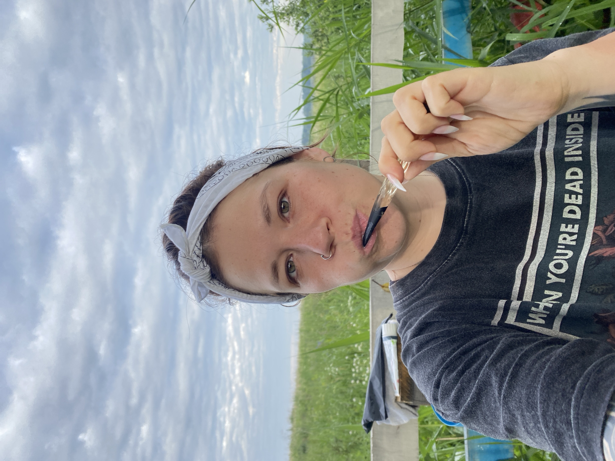 A selfie-style medium shot shows a woman holding a smoking pipe to her mouth, set against a backdrop of tall green grass and a cloudy sky. The woman, who appears to be outdoors, is wearing a dark t-shirt with a graphic print. A white bandana with a black pattern is tied around her head. She has a nose ring and hoop earrings. In the background, there is a white fence and some white objects, possibly bags or containers. The overall lighting suggests it is daytime, likely overcast. No specific location in Абзаново, Russia can be determined from the image.
