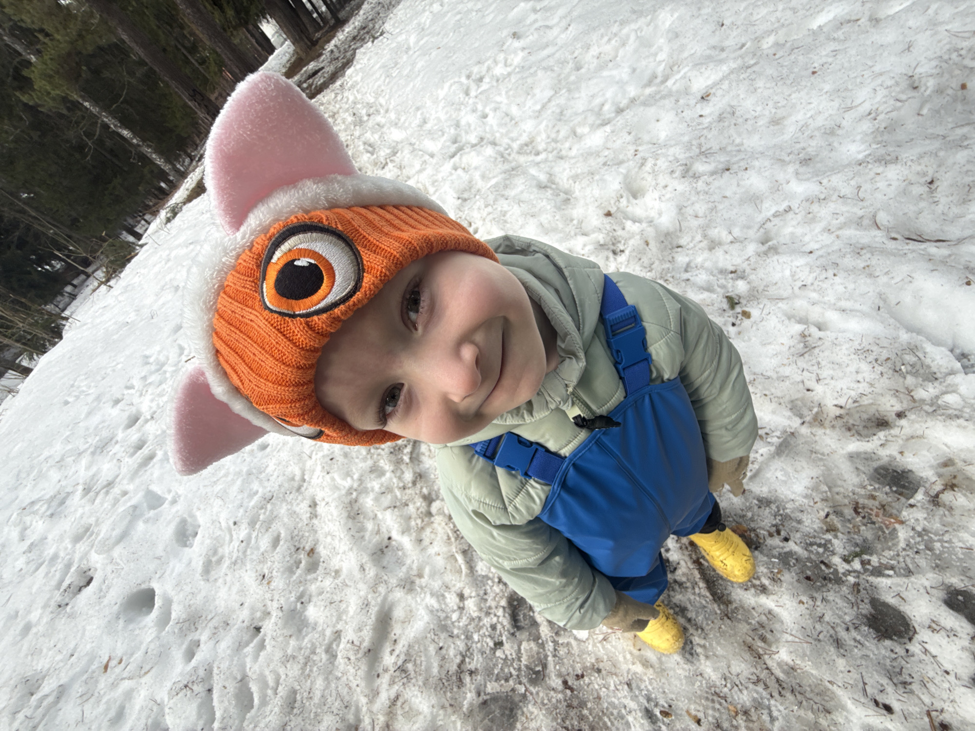 A child stands in a snow-covered area. The child wears a light green jacket, blue overalls, yellow boots, and an orange hat with a white trim, pink ears, and an embroidered eye design. The ground is covered in snow, with some visible dirt and debris. Trees are visible in the background. The location is possibly in a park or wooded area of Chaikovsky, Russia.