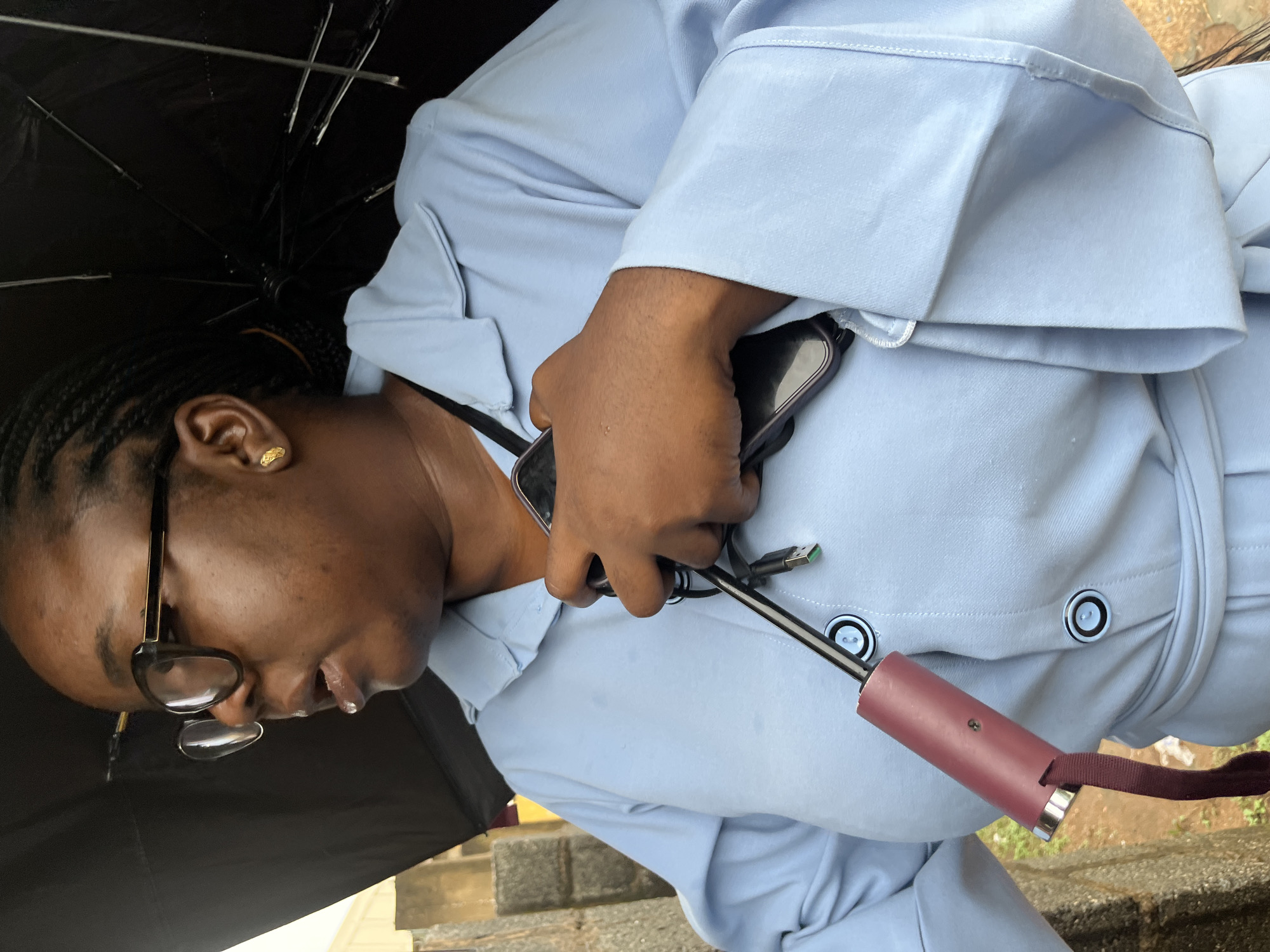 An outdoor, medium close-up shot shows a Black woman in a light blue collared dress. She is wearing black-rimmed glasses and a gold earring in her right ear. Her dark hair is styled in braids. She is holding a black umbrella with a red handle and a smartphone in her right hand, across her chest. The woman is looking to her left. The background shows a textured stone wall on the left and a blurred outdoor environment on the right. The lighting suggests it is daytime.