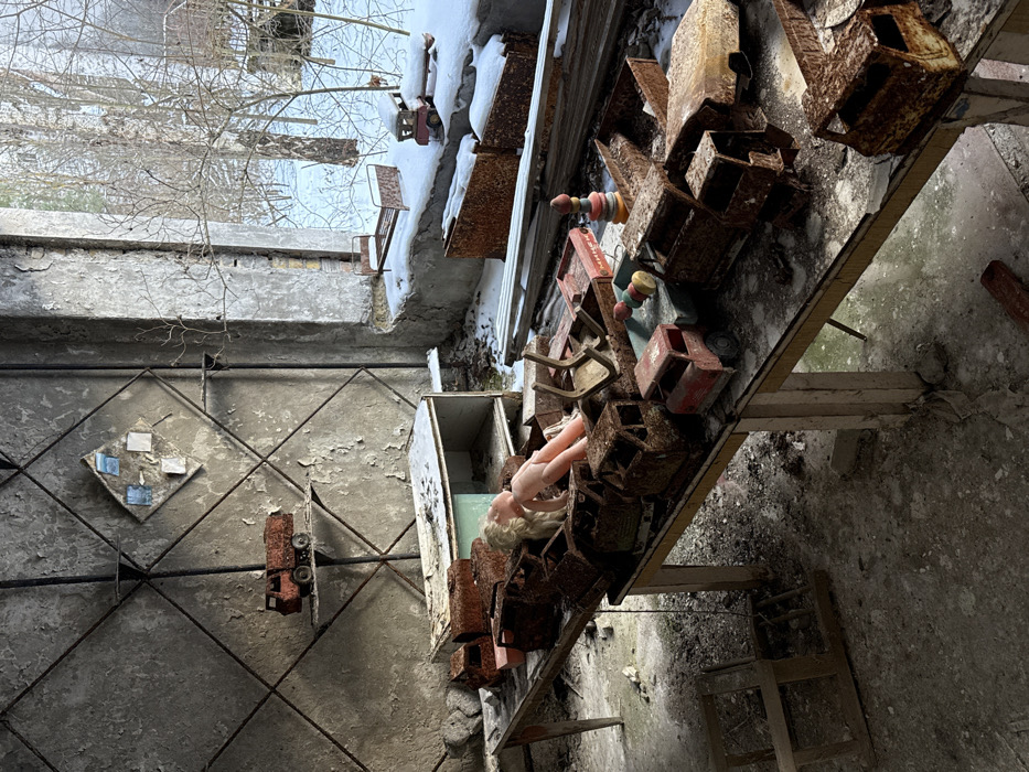 The image portrays an abandoned room in the city of Prypiat, Ukraine, during a winter season. The room's interior exhibits severe degradation, with peeling paint and plaster covering the wall, which features a prominent diagonal grid pattern. A large window occupies the right side, revealing a snow-covered exterior with bare, deciduous trees under an overcast sky. Snow has accumulated on the interior windowsill.
On the windowsill, a small rusted metal toy bed and a rusted toy truck are visible. The central focus of the room is a rectangular wooden table, its edges painted light yellow, covered with numerous rusted metal toy vehicles, predominantly trucks, in varying states of decay. A plastic doll with blonde hair and a pink torso lies face-up amidst these rusted toys. A colorful, multi-piece wooden stacking toy is also present on the table's right side.
Against the rear wall, a small metal shelf holds a single rusted toy truck. Above this shelf, a diamond-shaped panel featuring smaller square elements is affixed to the wall. Below the wall shelf, a low, open wooden cabinet shows signs of deterioration. The floor, appearing to be concrete, is covered with dust and debris. No individuals are present in the scene.