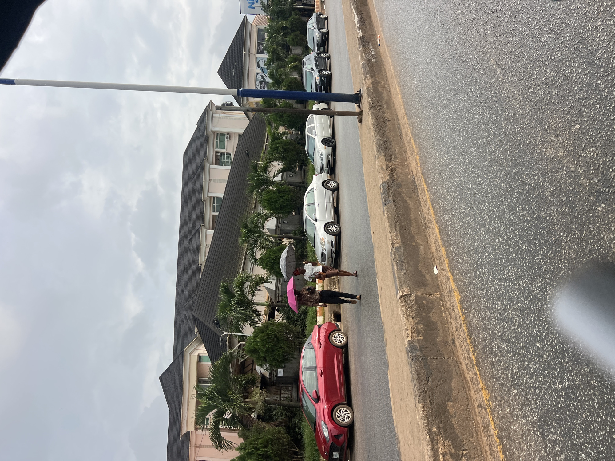 A low-angle, medium shot captures a street scene in Okpanam, Nigeria, under an overcast sky. In the foreground, a textured asphalt road with a yellow line marking the edge of a raised concrete curb dominates the lower half of the frame. Beyond the curb, a line of parked cars is visible, including a red sedan on the left, followed by several silver sedans and SUVs. Two individuals, one holding a pink umbrella and the other a black one, walk on the sidewalk near the red car, suggesting it may be raining. Behind the cars, a light-colored, multi-story building with a dark tiled roof and prominent architectural features is visible, flanked by lush green palm trees and other foliage. A tall, blue and white pole stands on the right side of the frame, partially obscuring the building and sky. The overall atmosphere is subdued, likely due to the cloudy weather.