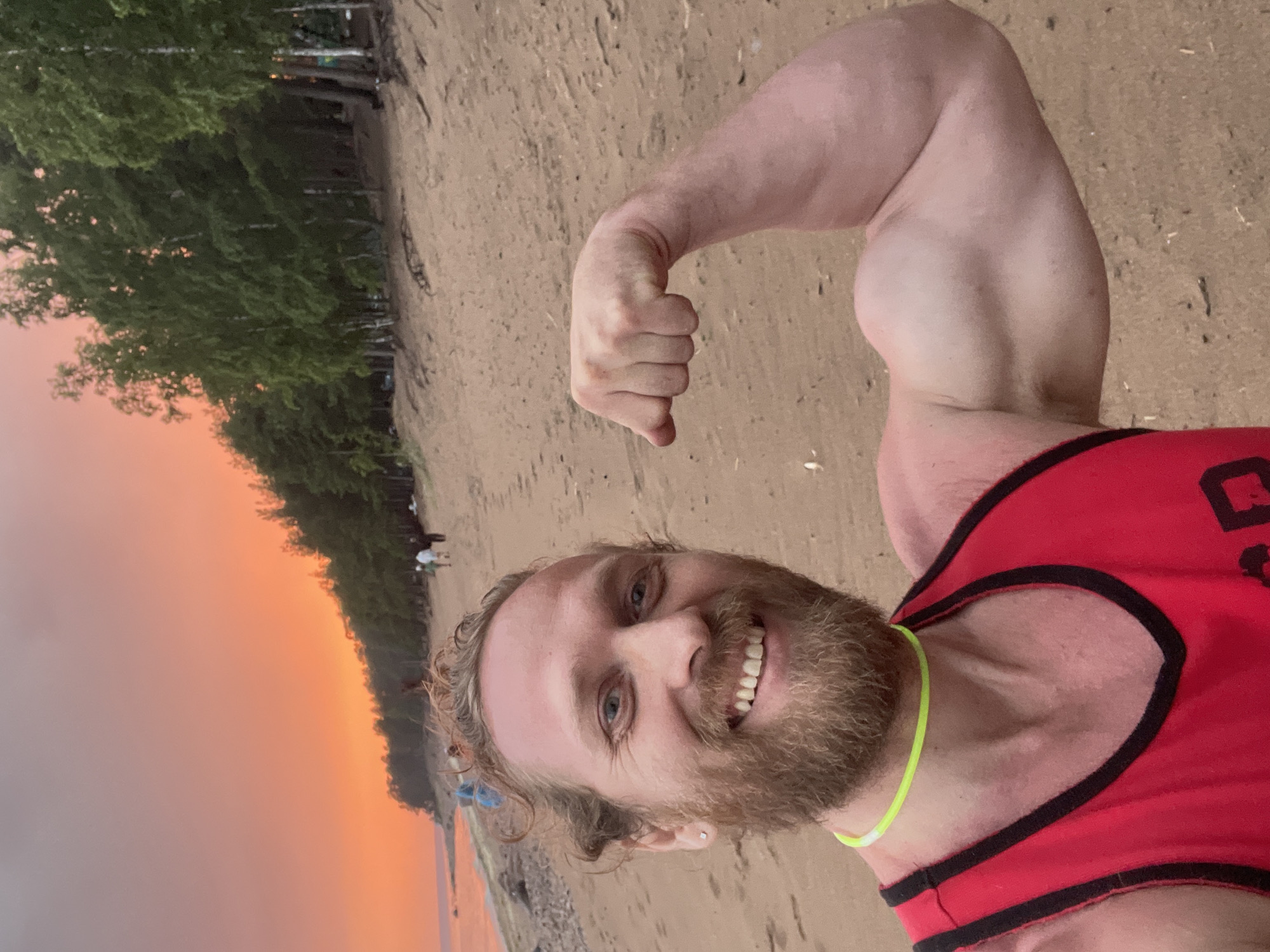 In Luzhki, Russia, a man with a beard and blond hair stands on a sandy beach at sunset. He is wearing a red tank top and a neon green necklace. He is flexing his bicep for the camera. In the background, there are trees and a body of water. The sky is a vibrant orange and pink. Several other people are visible in the distance, walking along the beach.