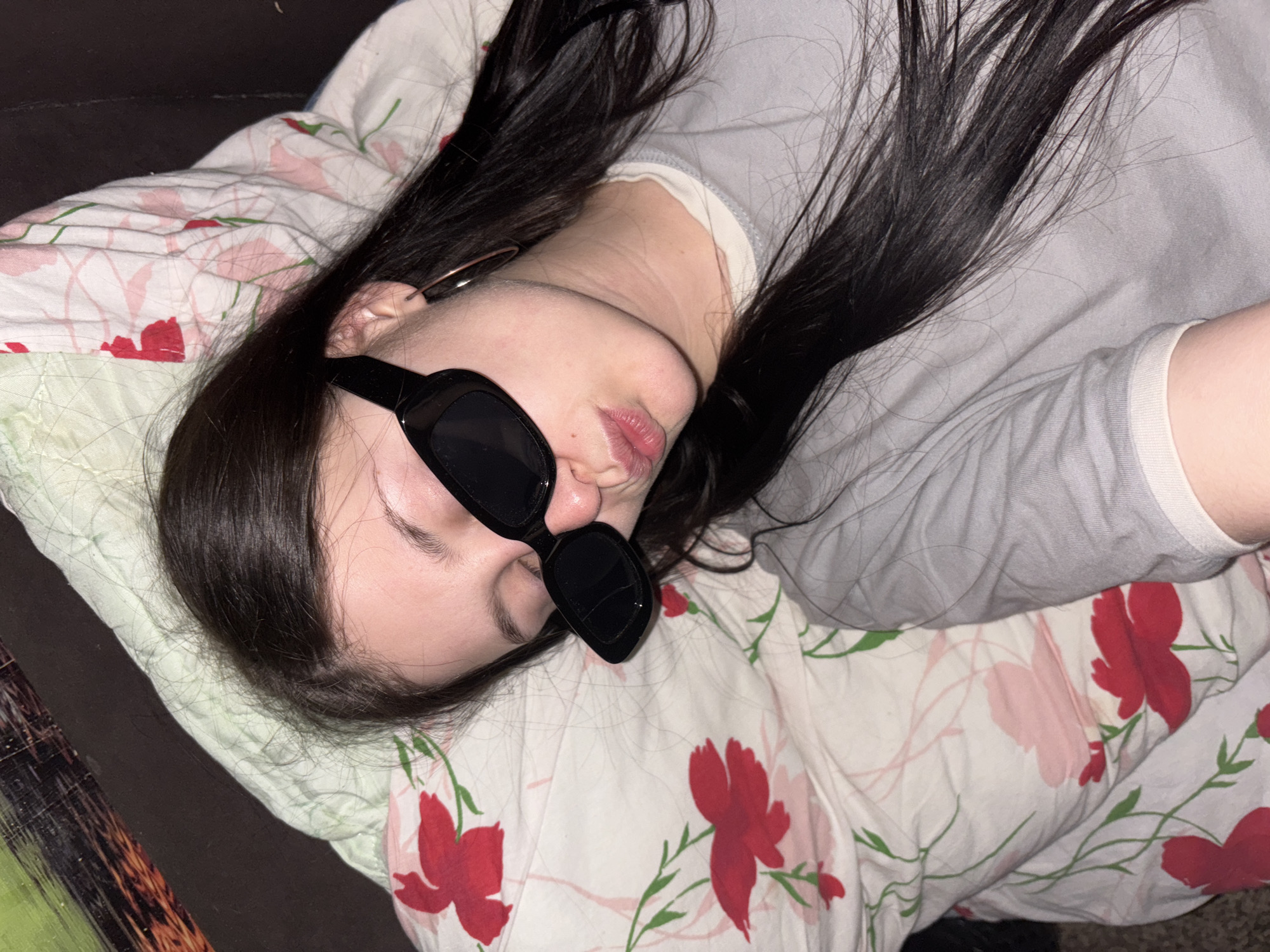 A person is lying on a pillow with a floral pattern, wearing dark sunglasses and a light-colored shirt. Their dark hair is spread around their head. The scene appears to be indoors, potentially in a domestic setting. The location is not visually discernible from the image.