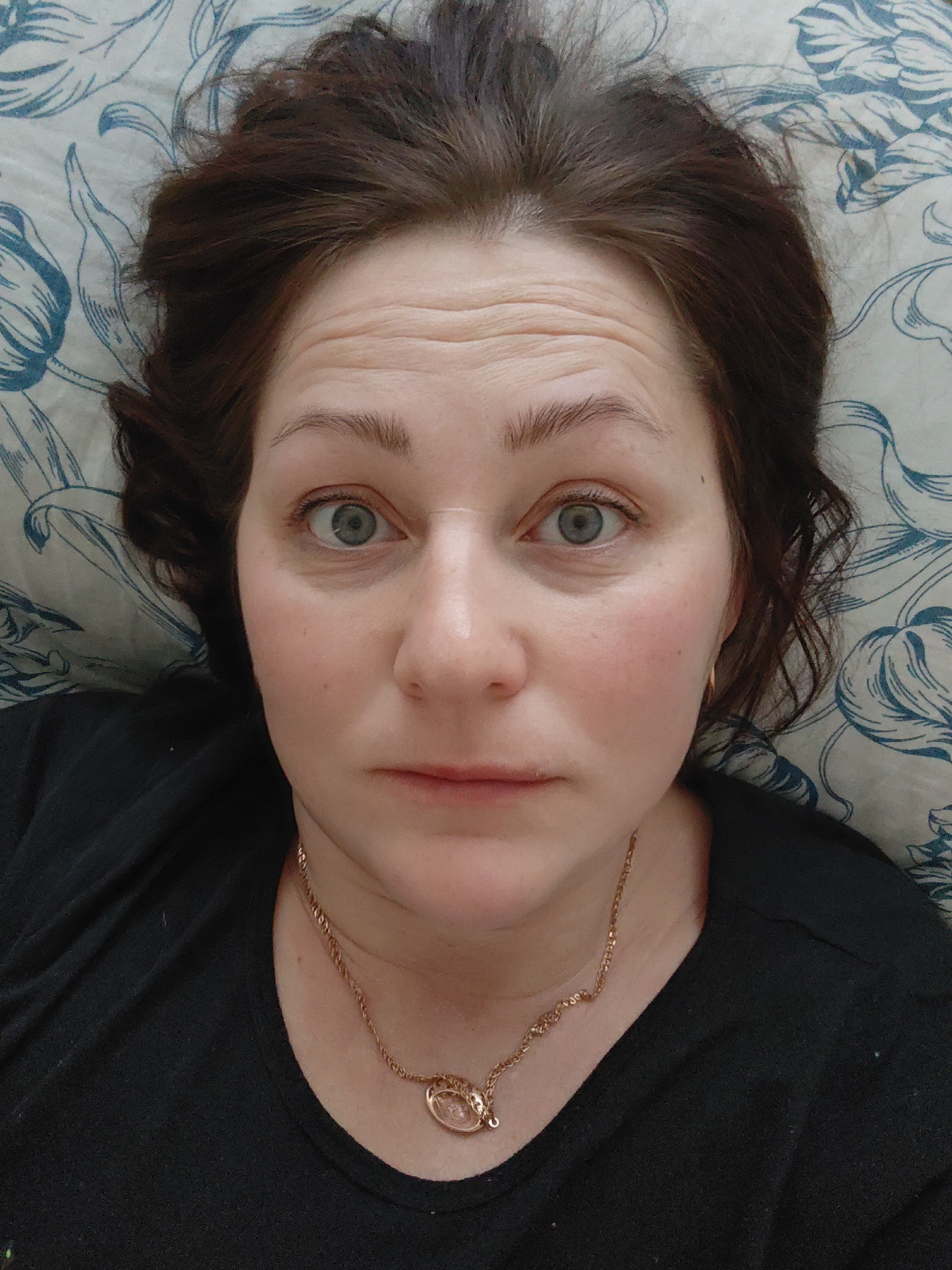 A close-up portrait shows a woman lying down, looking directly at the camera. Her head is resting on a light-colored pillow or bedding featuring a repeating pattern of stylized blue/teal botanical motifs. She has dark brown, slightly dishevelled hair that frames her face and spreads out on the patterned fabric. Her skin is fair, with a natural flush on her cheeks. Several horizontal lines are visible on her forehead, indicating raised eyebrows, which, combined with her wide, open blue/grey eyes, convey an attentive, focused, or slightly surprised expression. She is wearing a simple black top or shirt. Around her neck, she has a gold-toned chain necklace with a small, oval, translucent or light-colored pendant. The lighting is soft and even. The scene appears to be indoors, likely in a bedroom setting, in Markovskii, Russia. No visible text is present in the image.