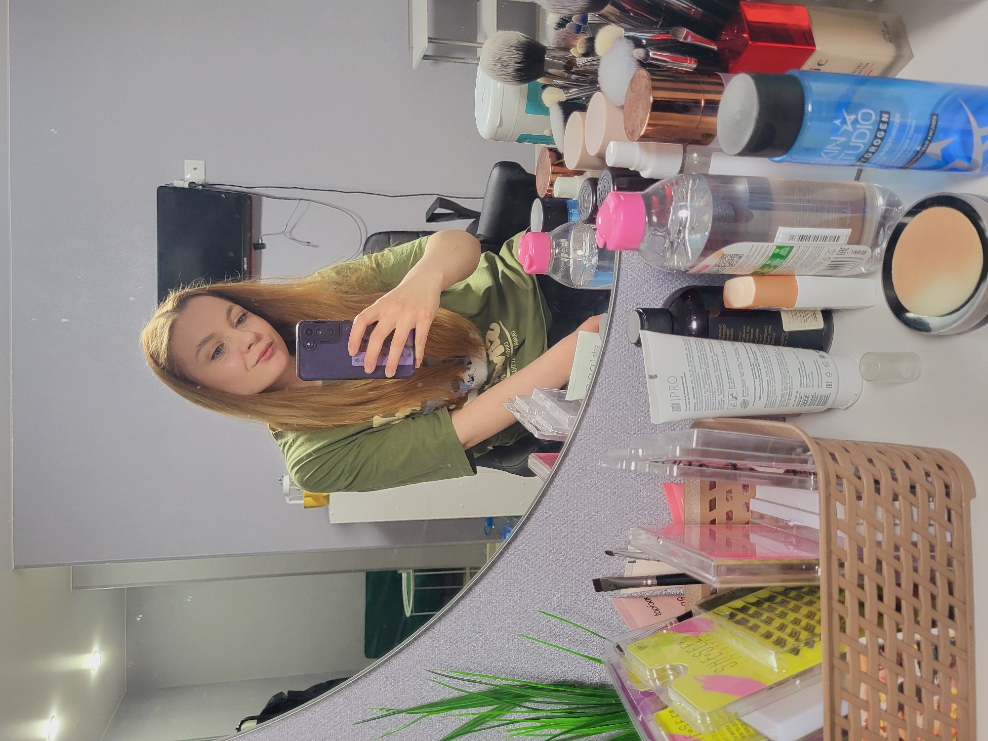 A woman is taking a selfie in a mirror. She is wearing a green t-shirt and has long brown hair. The background shows a collection of makeup and skincare products on a vanity or counter. Brands such as SKIN STUDIO HYDROGEN, TOPFACE, IPRO, and others are visible. There is also a basket containing more makeup items. In the reflection, part of a room is visible with a TV mounted on the wall. While not explicitly confirmed, this photo seems to be taken in Chaikovsky, Russia, based on the OCR text.