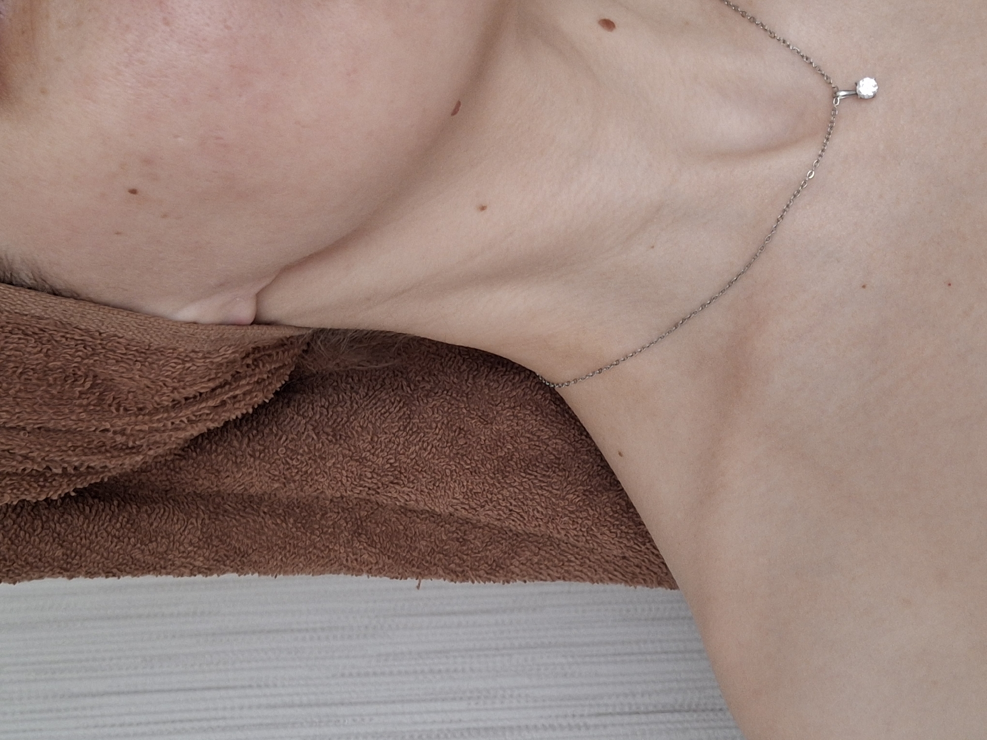 A close-up photograph depicts a person's head and neck, adorned with a silver necklace featuring a single, small diamond or crystal. The person's head rests on a brown towel, which is placed on a textured, white surface. The subject has several moles on their neck and face. The location, Chaikovsky, Russia, is not discernible from the visual information provided.