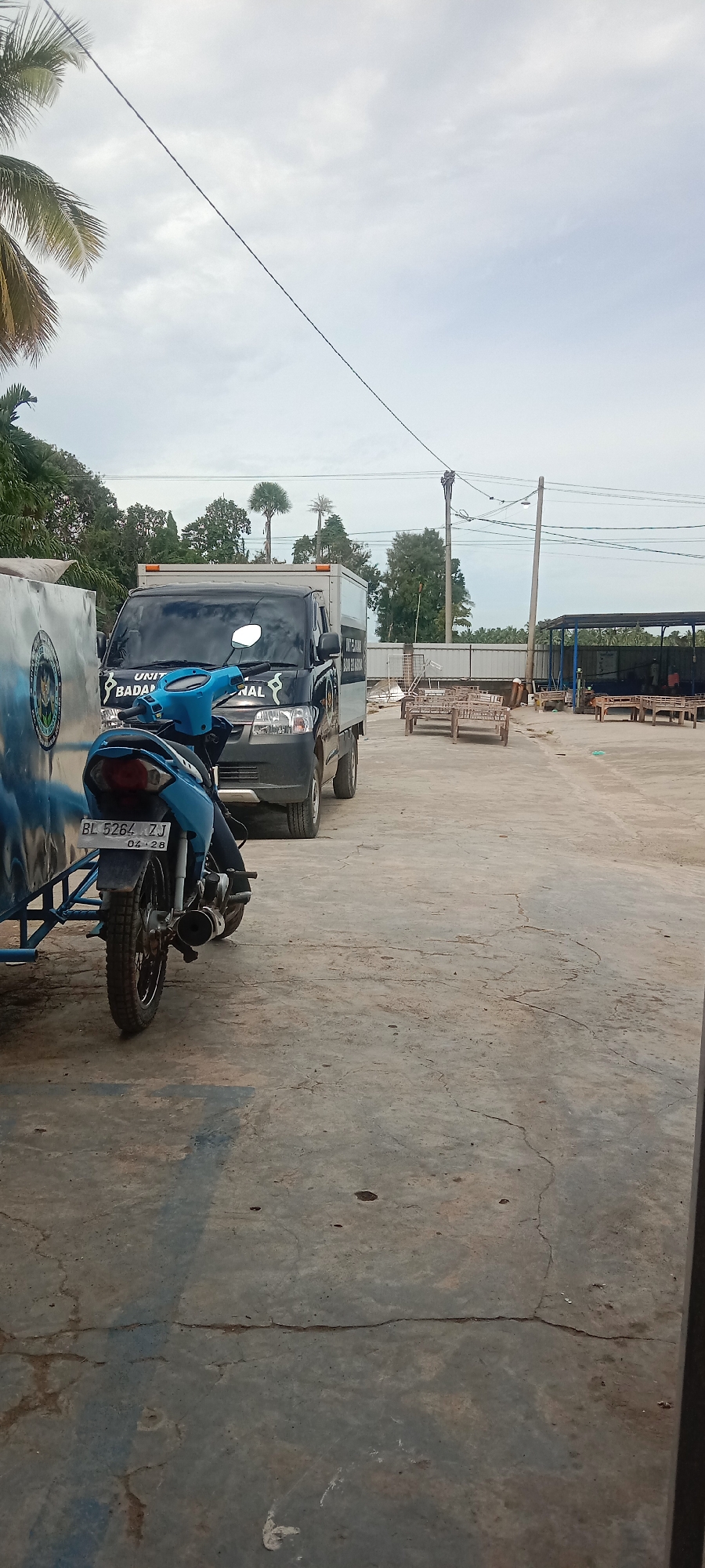 A motorcycle with a sidecar, a dark gray pickup truck, and a utility building are present in this outdoor scene. The motorcycle is blue and white and is parked in the foreground. A license plate on the motorcycle reads "BL 5264 ZJ." The pickup truck is parked behind the motorcycle. A sign on the side of the truck reads "UNIT BADAN NASIONAL." A utility building with a corrugated metal roof is visible in the background, along with trees and power lines. A person is seen working on the power lines in the distance. The ground is paved with concrete.