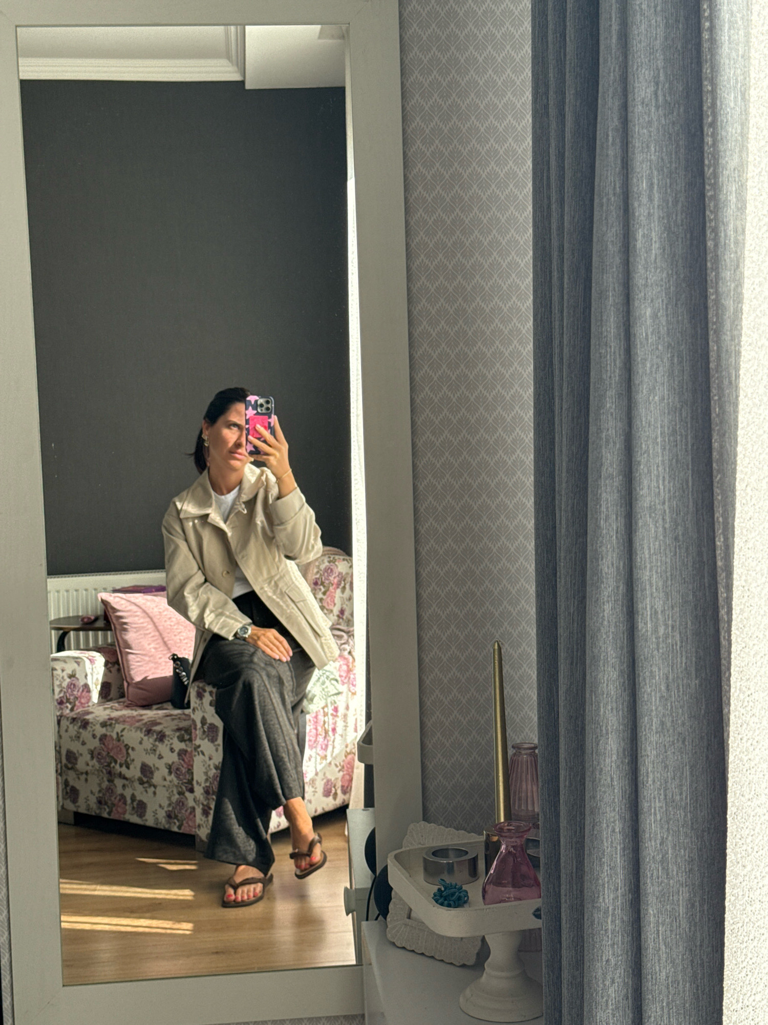 The image shows a woman in her late 20s or early 30s taking a mirror selfie in what appears to be a bedroom or dressing area in Tbilisi, Georgia. She is seated on a floral-patterned sofa, wearing a light-colored trench coat over a white t-shirt and dark-colored wide-leg pants. She has dark hair pulled back and is wearing hoop earrings and a wristwatch. Her right hand is holding a pink and black floral-patterned smartphone to take the picture. Her feet are bare, with flip-flops placed nearby.
The room features dark gray walls in the background, a white radiator, and a large white-framed mirror. To the right of the mirror, a sheer gray curtain hangs over a window with textured white blinds. A small decorative table with items like a golden candle, pink glass vases, and a white stand with a teal decorative item are visible. The floor is made of light-colored wood, reflecting sunlight coming from the window. The sofa is upholstered in a chintz fabric with a floral pattern of pink roses and green leaves. A pink throw pillow rests on the sofa. The overall impression is of a well-lit, contemporary living space. The presence of a radiator suggests it could be a cooler season. The style of the furniture and decor is consistent with modern apartments found in Tbilisi.