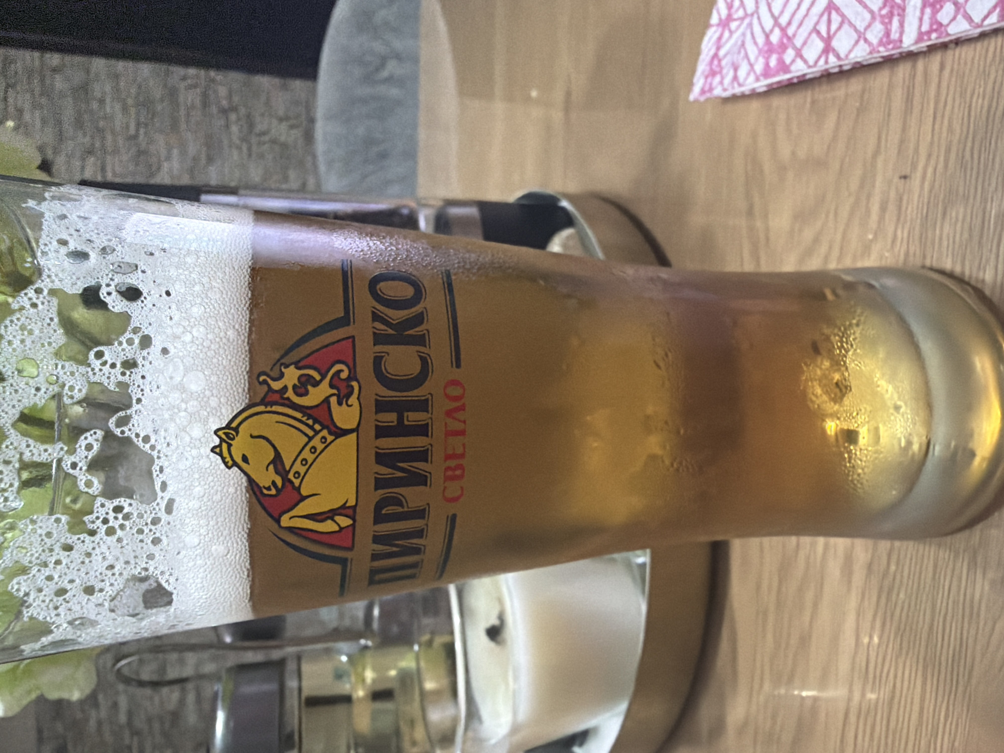 The image displays a tall, frosted glass of light-colored beer, likely a lager, resting on a light wooden table. The glass has a significant head of white foam, and condensation is visible on its surface, indicating the beer is cold.
The main subject of the image is the beer glass, which features the brand name "ПИРИНСКО" (PIRINSKO) prominently displayed in black letters with a gold outline. Below the brand name, the word "СВЕТЛО" (SVETLO), meaning "Light," is written in red Cyrillic script, indicating the type of beer. A distinctive logo is visible on the glass, depicting a stylized yellow horse or stallion on a red background with a black outline.
The setting appears to be indoors, possibly a bar or restaurant in Shumen, Bulgaria, aligning with the Bulgarian text on the beer glass. A pink and white patterned napkin is partially visible in the upper right corner of the frame. Underneath the glass, a metallic or silver tray/coaster is discernible. The background is softly blurred, showing hints of dark objects on the left and a lighter, perhaps cushioned, surface on the right, along with more of the wooden table texture. No people are visible in the image, and no specific activity beyond the presence of the beer is depicted. The lighting suggests an indoor ambiance without indicating a specific time of day.