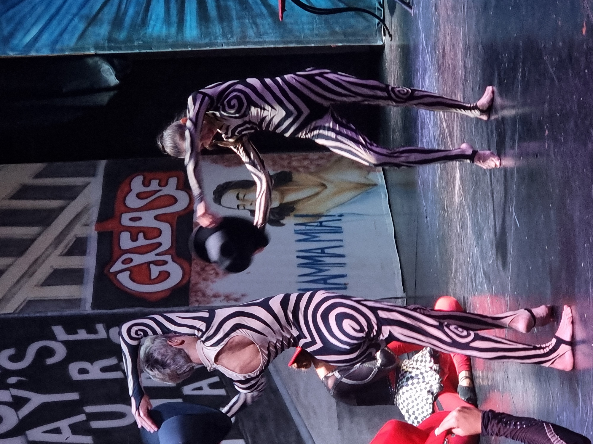 Two performers in black and white spiral-patterned bodysuits are on a stage. The performer on the left is facing away from the camera, with her left arm raised and holding a black hat. The performer on the right is facing left, with both arms extended, also holding a black hat. Behind them, a backdrop features large white letters spelling "AYS URE" on the left and the word "GREASE" in red and white lettering on the right. Another banner behind the performers reads "MAMMA MIA!" in blue spray-paint-like letters. The stage floor is dark and reflective. The scene appears to be a theatrical performance. This image was taken in Manavgat, Turkey.