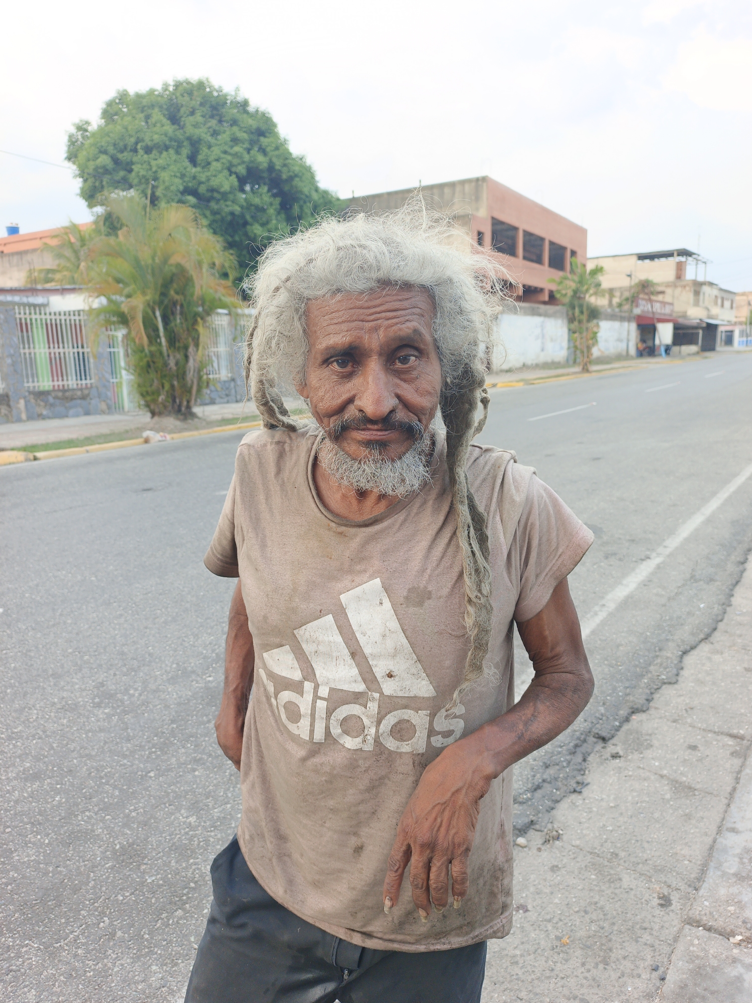 An elderly man stands on the side of a road in Tinaquillo, Venezuela. He has long, matted grey dreadlocks and a grey beard. He is wearing a dirty, light brown Adidas t-shirt and dark pants. The man is facing forward, looking at the camera with a neutral expression. The road is cracked and has some debris. Buildings, a fence, and trees are in the background. The sky is overcast.
