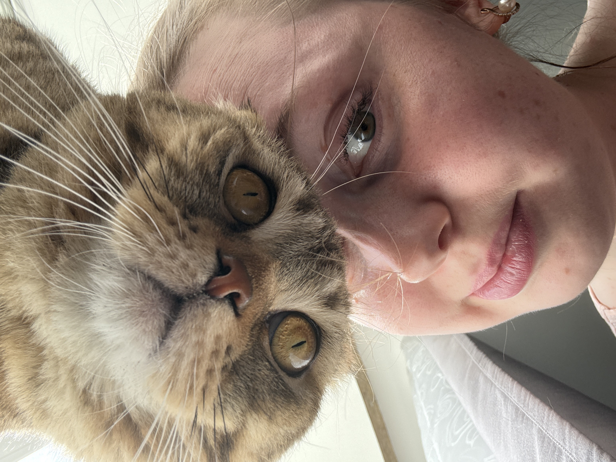 A close-up selfie image features a blonde woman and a brown tabby cat. The woman is fair-skinned with freckles, and her face is partially obscured by the cat. Her lips are pink, and her eyes are green. A small earring is visible. The cat's face takes up a large portion of the left side of the image, with its amber eyes and numerous white whiskers being prominent features. The image is taken from a high angle, looking down at the subjects. The background is out of focus but appears to be indoors, with hints of a white surface and grey fabric. The location and city are unknown.