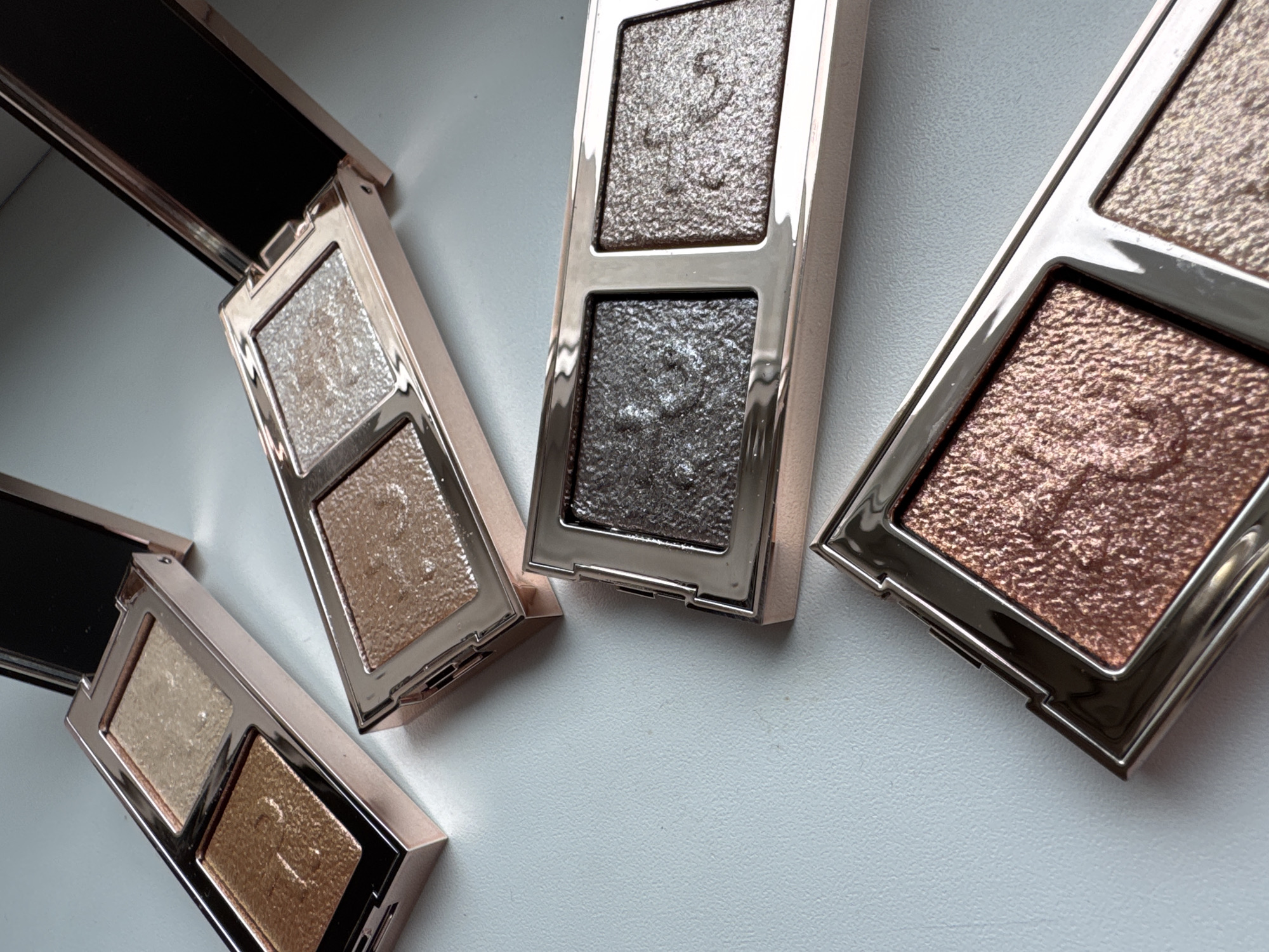 The image shows three open, two-pan eyeshadow palettes and one closed eyeshadow palette, arranged on a light-colored surface. The open palettes display various shades of glittery eyeshadows. The palettes are likely indoors and may be used for cosmetic purposes. The specific location within Ждановіцкі сельскі Савет, Belarus cannot be determined from the image.
