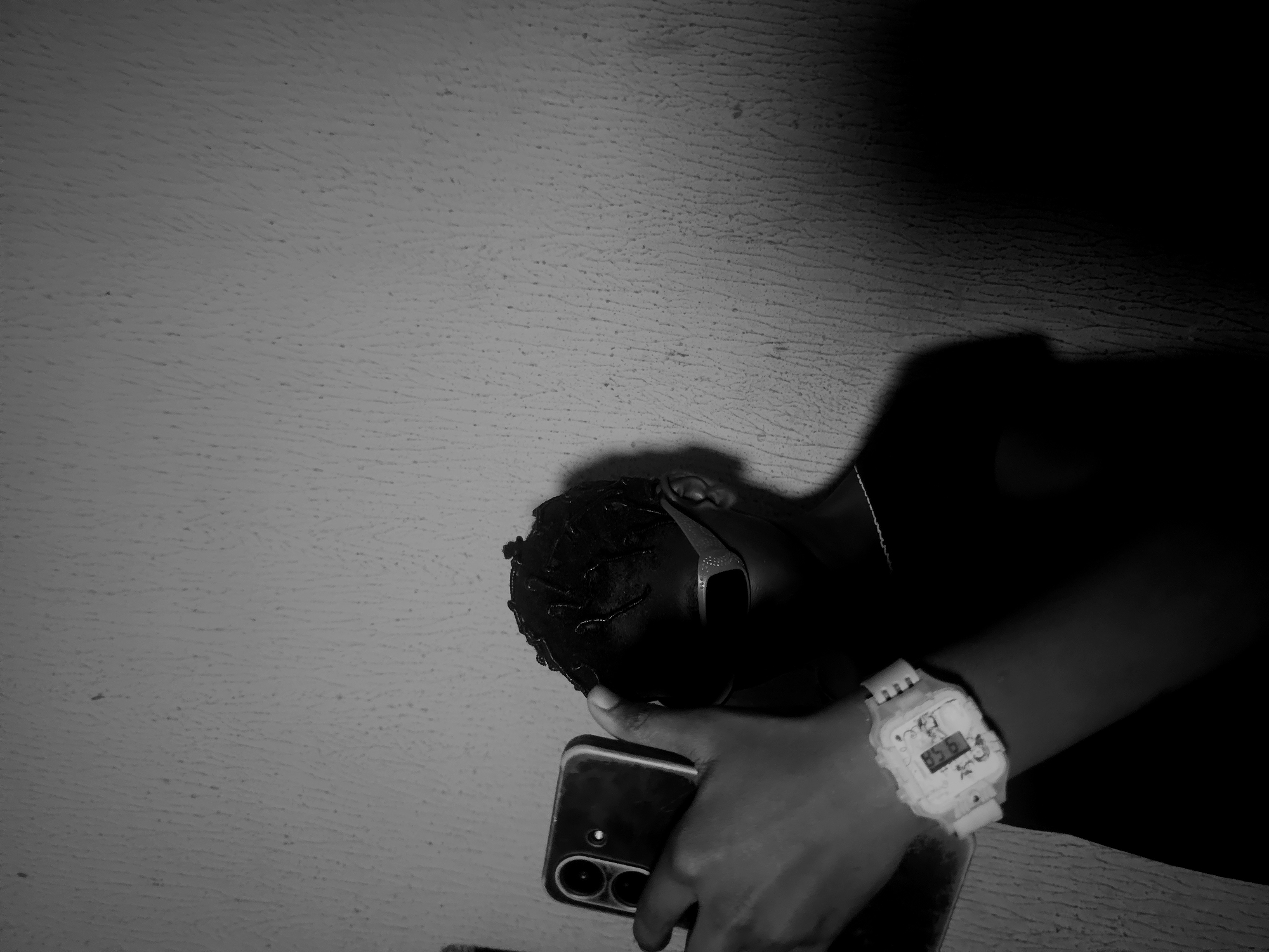 A low-angle, black and white photograph depicts a person holding a smartphone. The person is wearing sunglasses and a white watch. The background appears to be a textured wall. The location cannot be determined.