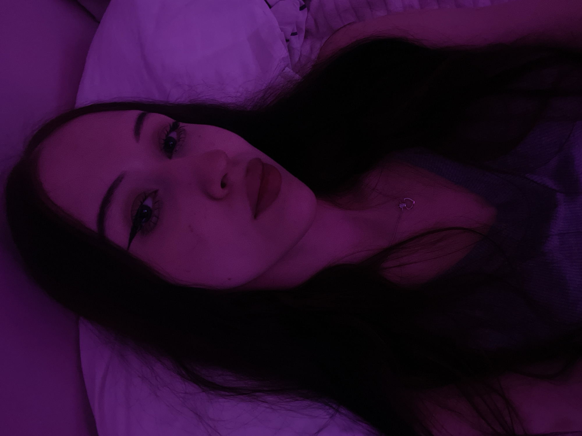 In this close-up, indoor selfie, a young woman with long, dark hair is shown with her head tilted slightly. She is lying on a pillow, illuminated by a purple light, which casts a strong hue across the scene. Her makeup is noticeable, with winged eyeliner and dark lipstick. She is wearing a grey or purple top, and a small, heart-shaped pendant necklace is visible. The background appears to be a bed with a pillow and a hint of a white sheet or blanket. There is no indication of the city of Votkinsk, Russia, in the image.