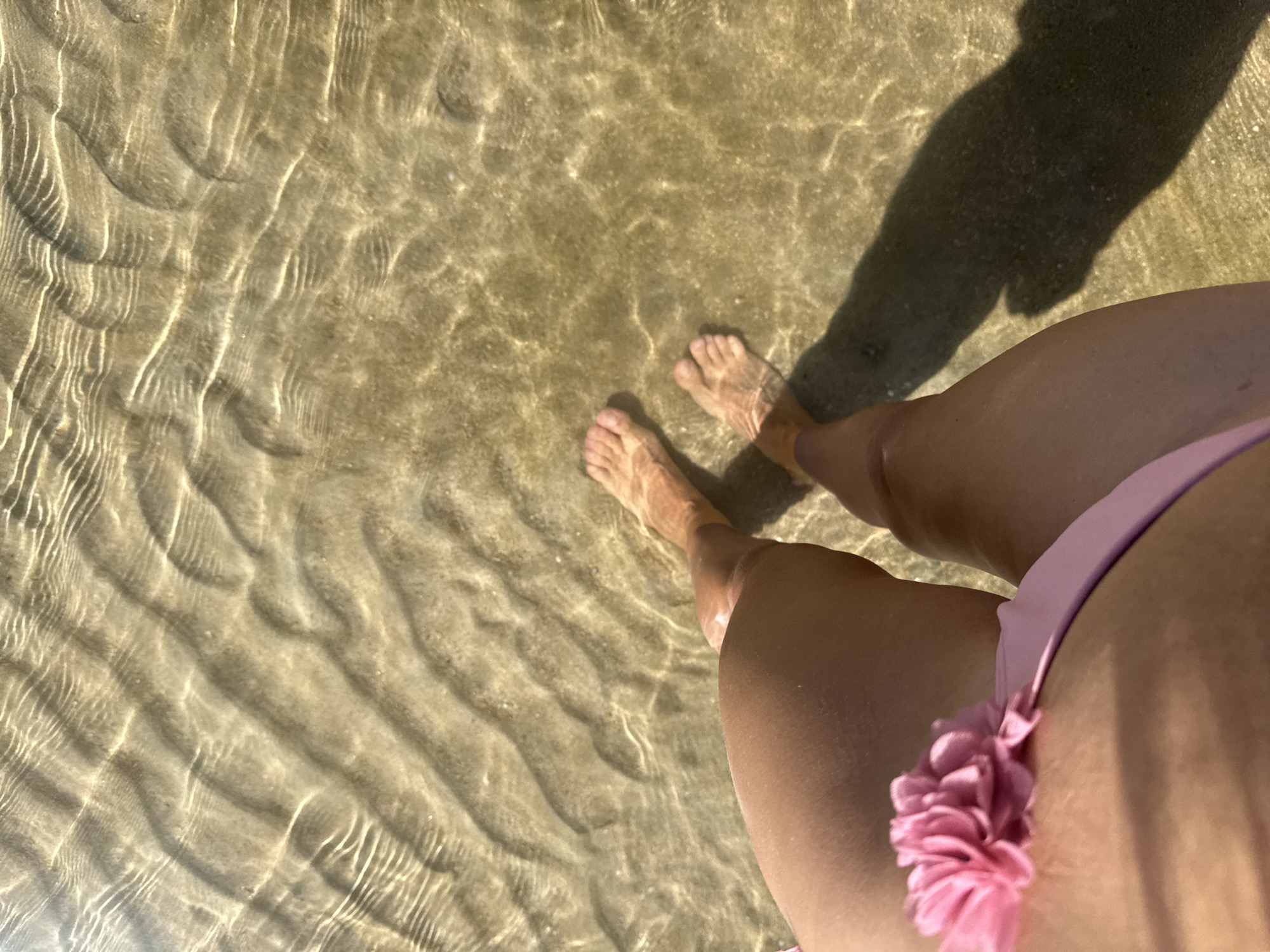 This is a top-down, medium shot of a person's legs and feet standing in shallow, clear water at a beach in Baan Chaweng Noi, Thailand. The sand beneath the water has visible rippling patterns. The person is wearing a pink bikini with a floral embellishment on the side. The water is shallow enough that the feet are partially submerged. A shadow of the person is cast on the sandy seabed. The lighting suggests it is daytime.