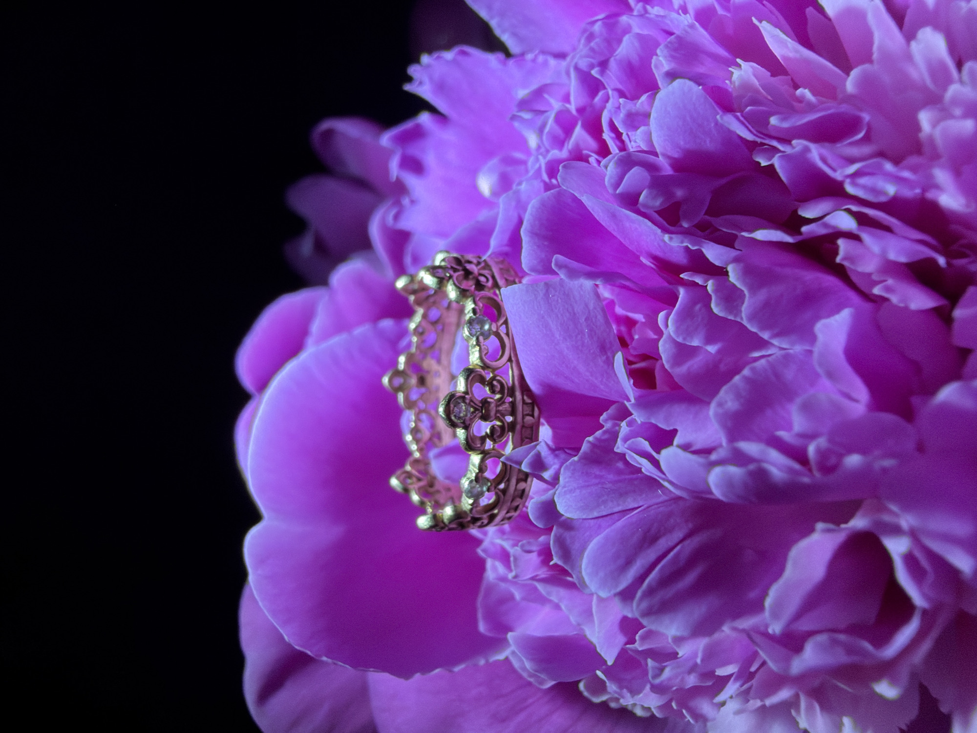 The image shows a gold ring with a floral design and small stones resting on the petals of a bright pink peony flower. The background is dark and out of focus. There is no evidence of the image being taken in Chaikovsky, Russia.