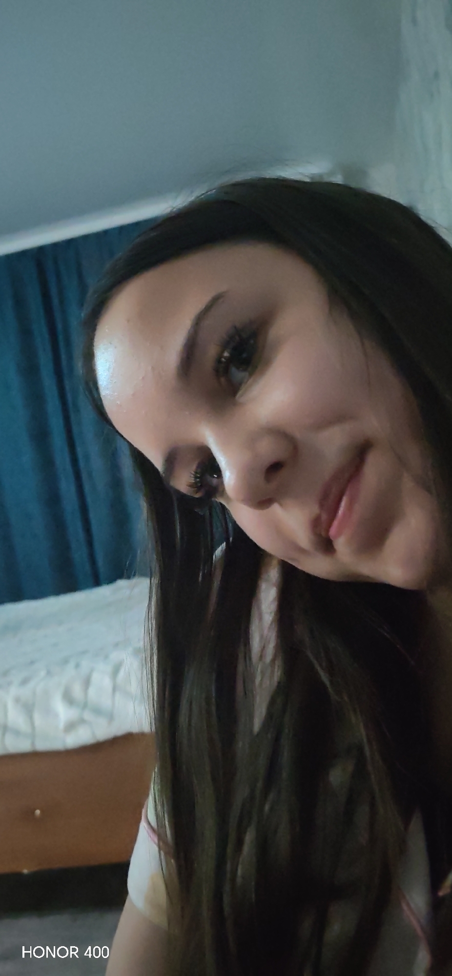 A close-up portrait shows a woman with long, dark hair, fair skin, and light-colored eyes, smiling slightly. She is indoors, with a bed visible in the background, covered with a white, textured bedspread. Behind the bed are dark blue curtains. The lower left corner of the image contains the text "HONOR 400". While the location provided is Noviy, Russia, there is no specific information in the image to definitively confirm this location.
