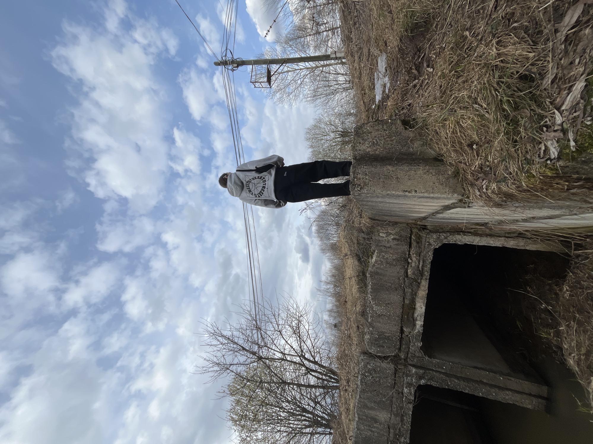 The image shows a lone person standing on a concrete structure, viewed from behind, in an outdoor setting in Sosnovo, Russia. The individual is wearing a light grey hooded sweatshirt and dark pants. The back of the hoodie features a prominent circular graphic design with text, including "THE REVOLUTIONARY PROJECT" and "ECOLOGY" in English. Other symbols and text within the design are less clear.
The person is positioned on a rugged concrete surface, which appears to be part of a bridge or culvert, given the large, dark, square openings visible beneath it. The surrounding environment consists of dry, brown grass and sparse vegetation, suggesting late winter or early spring. Several bare deciduous trees are scattered across the landscape in the background. A wooden utility pole with multiple power lines is visible on the left side of the frame, contrasting with the natural elements. The sky above is bright blue with scattered white cumulus clouds, indicating daytime. The scene evokes a sense of quiet observation in a semi-rural or industrial area.