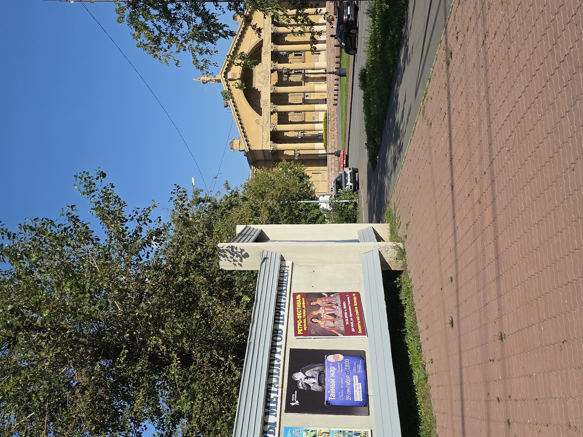 The image depicts a street scene in Chelyabinsk, Russia, featuring a cultural institution and its surroundings on a clear, sunny day. In the foreground, a red brick sidewalk stretches across the bottom of the frame, bordered by a grassy strip. To the left, a concrete wall displays several posters, one advertising a performance titled "Тайный жар" (Secret Heat) by the "Театр в Потемках" (Theater in the Dark), scheduled for September 19th at 18:00 at the "Дк ЧМК" (DK ChMK). Another poster promotes a "Ретро-фестиваль" (Retro Festival) featuring music, dance, and a fashion show, scheduled for October 10-10, 2020, at the "ДК ЧМК" on prospekt Gagarina, with tickets available on kassy.ru.
Dominating the background is a large, yellow, neoclassical-style building with a grand facade featuring columns and an arched entablature, topped with a golden statue. This building is likely the "ДК ЧМК" (House of Culture of the Chelyabinsk Metallurgical Plant), a prominent cultural center in Chelyabinsk. A street runs in front of the building, with a few cars visible: a white SUV, a red sedan, and a dark-colored hatchback. Trees with green foliage are present on the left and right sides of the image, partially obscuring the view of the building and the sky. Overhead, telephone wires crisscross the blue sky. The overall atmosphere is one of civic engagement and cultural activity within an urban setting.