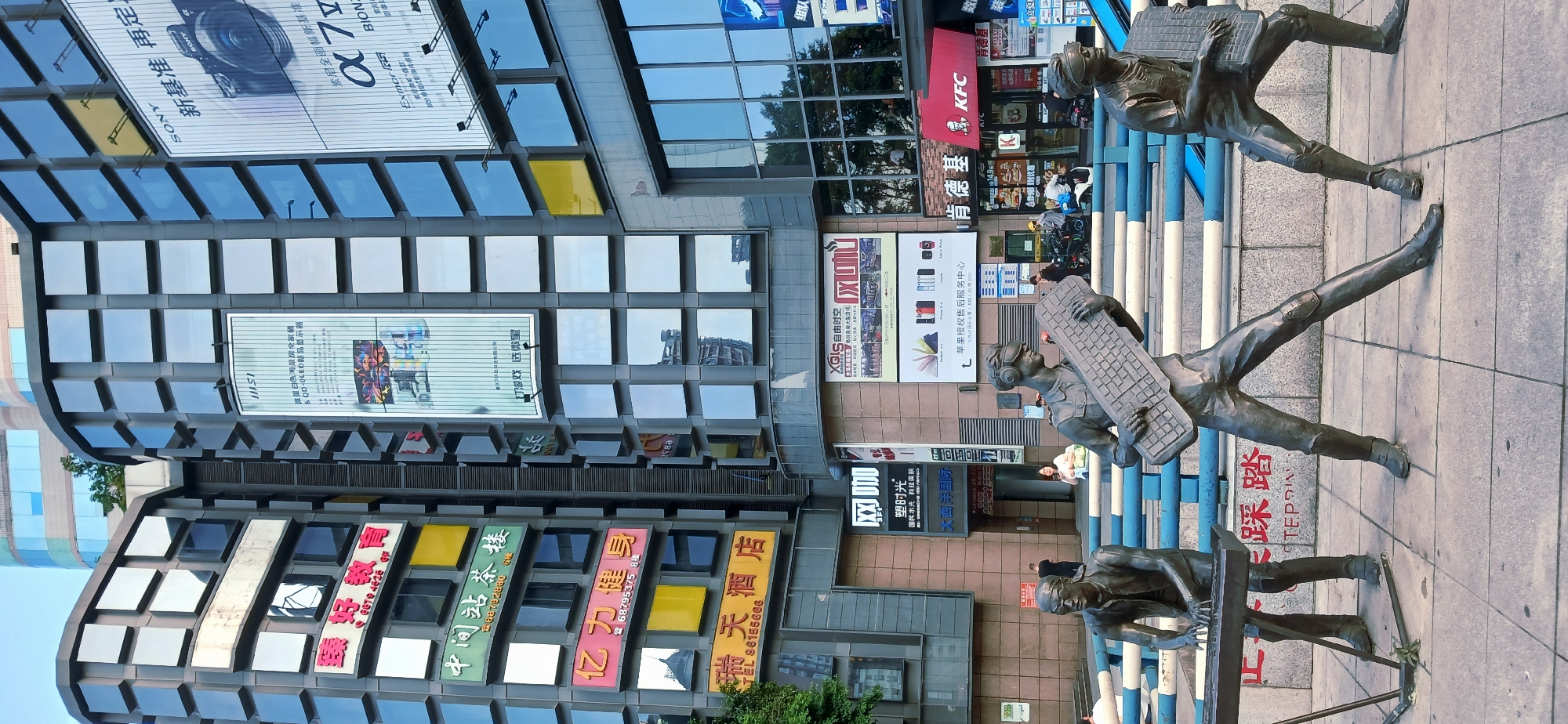 The image depicts a street scene in Jiulongpo District, China, featuring three bronze statues of figures working at keyboards, installed on a sidewalk. Behind the statues is a multi-story building adorned with various advertisements. These ads include those for Sony cameras, MSI products, and a "KFC" restaurant. Also noticeable are Chinese characters indicating other businesses, such as a tea house and a hotel. The architectural style of the building includes a grid pattern of windows. The statues are positioned adjacent to a blue and white striped barrier. The foreground shows the weathered surface of the sidewalk. The image is taken from a low angle, emphasizing the height of the building and the size of the statues.