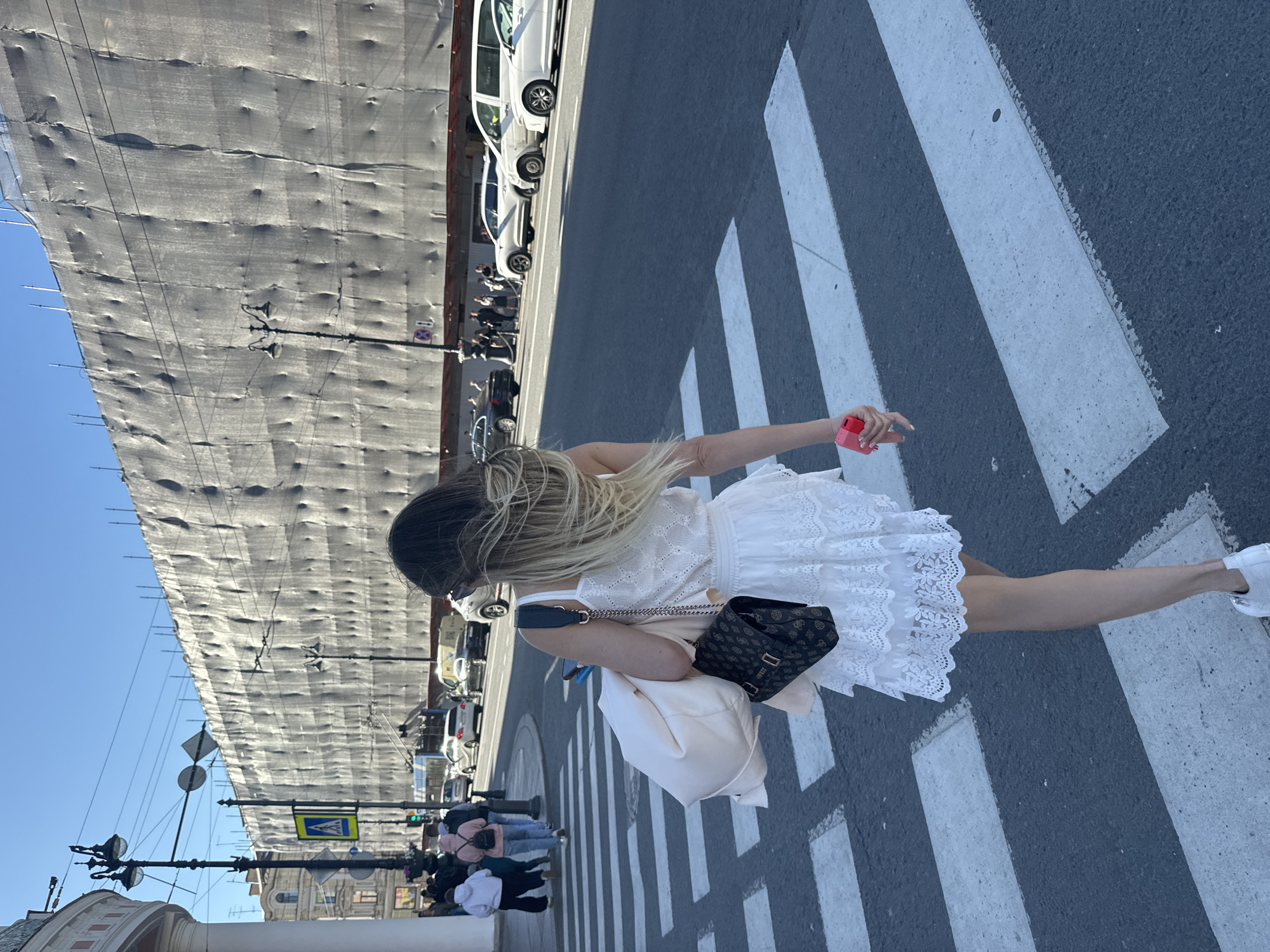 The image depicts a woman in a white lace dress walking across a crosswalk on a sunny day in Saint Petersburg, Russia. She is carrying a cream-colored jacket over her left arm and a dark brown patterned purse with gold accents on her right shoulder. In her right hand, she holds a bright pink object, possibly a phone. Several cars are visible on the street, and a large building covered in grey scaffolding or sheeting is in the background. Pedestrians can be seen on the sidewalk to the left. The sky is clear and blue.
