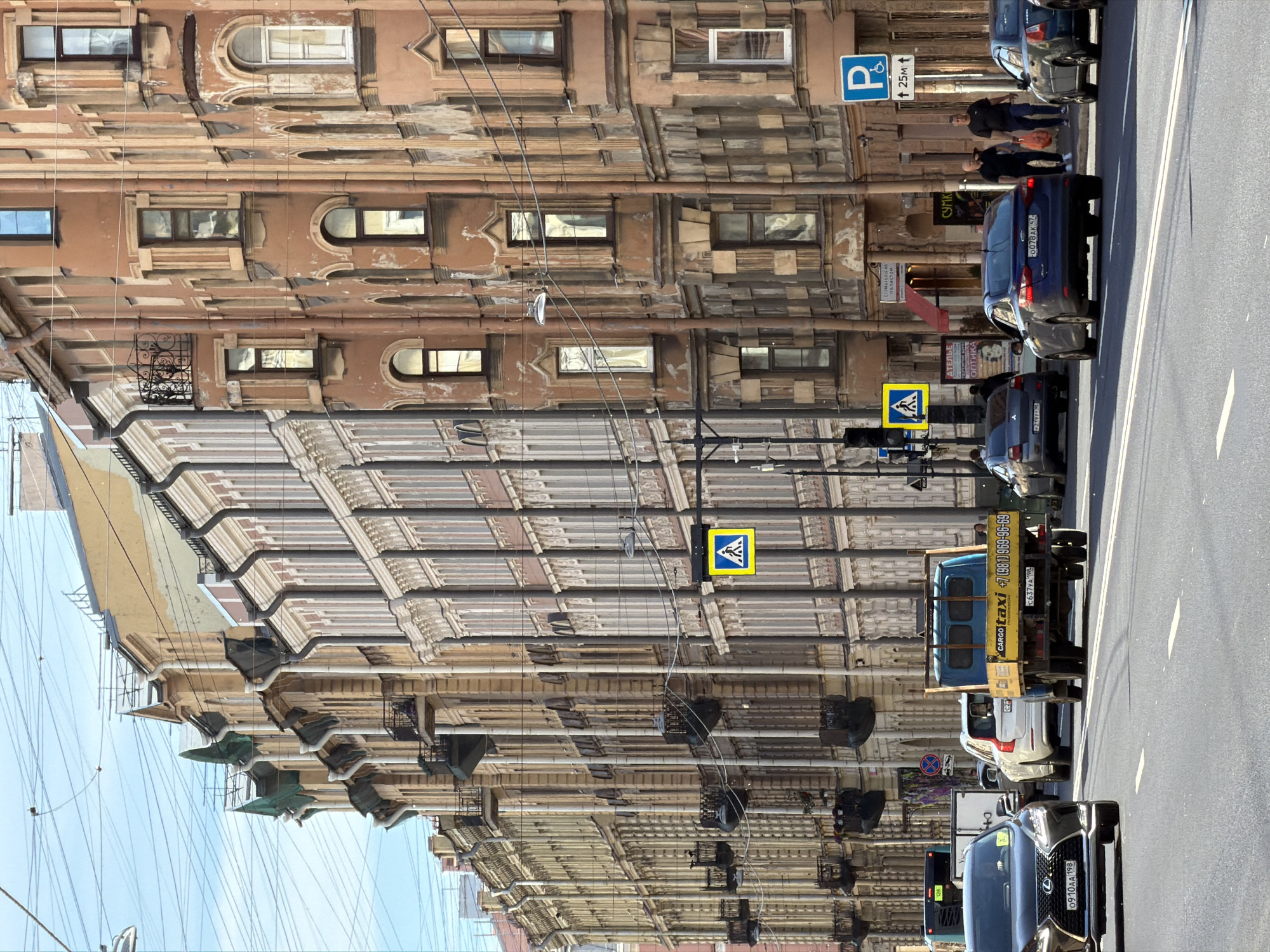 This is a street-level, eye-level, slightly tilted-down shot of a street in Saint Petersburg, Russia. The scene is dominated by the ornate architecture of old buildings lining the street, with their numerous windows, decorative facades, and intricate moldings. The buildings appear to be made of weathered, pale brown material, and many of them show signs of age.
A line of cars is parked or moving along the right side of the street. Visible vehicles include a blue sedan with a license plate ending in "AK 57", a white sedan, and a blue truck with "CARGO taxi" written on its side, along with a phone number. Further down the road, a dark SUV with a license plate ending in "AA 198" is visible. There are also pedestrian crossing signs with yellow diamonds and black pedestrian silhouettes.
Several people are visible on the sidewalk, mostly dressed in dark clothing. One person is carrying an orange bag. The sky is a clear blue, suggesting fair weather, and the lighting indicates it is daytime, likely late morning or early afternoon, with the sun casting shadows on the street and buildings.
There are various signs visible on the buildings, including one with Cyrillic text that appears to be for "ОПТИКА" (Optics) and "СТОМАТОЛОГИЯ" (Dentistry). A blue parking sign with a white "P" and an arrow is also present. The image captures a typical urban scene in a historic European city, with a mix of old architecture, modern vehicles, and pedestrian activity.