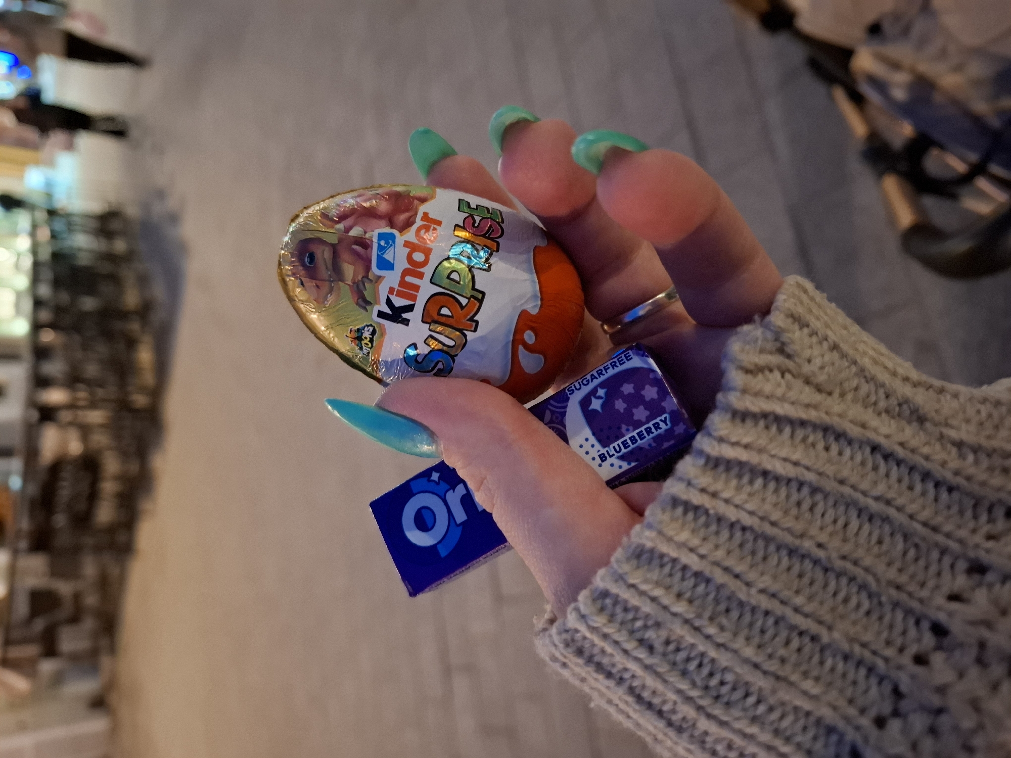 A person holds an assortment of treats in their hand against a blurred background.
The person has fair skin and manicured nails, with a turquoise polish on the index and pinky fingers, and a light green on the remaining fingers. The person is wearing a silver ring on their ring finger and a beige, knitted sleeve is visible.
The person holds:
* A Kinder Surprise egg, showing a cartoon dinosaur
* A small package of blueberry-flavored, sugar-free candy
* A small package of Oreo cookies
The background is blurred, appearing to be an outdoor urban environment at night. There are lights and indistinct shapes of buildings visible, resembling a street scene, although specific details are impossible to discern. No confirmed information is possible to determine about the location of the image in Novi Sad, Serbia.
