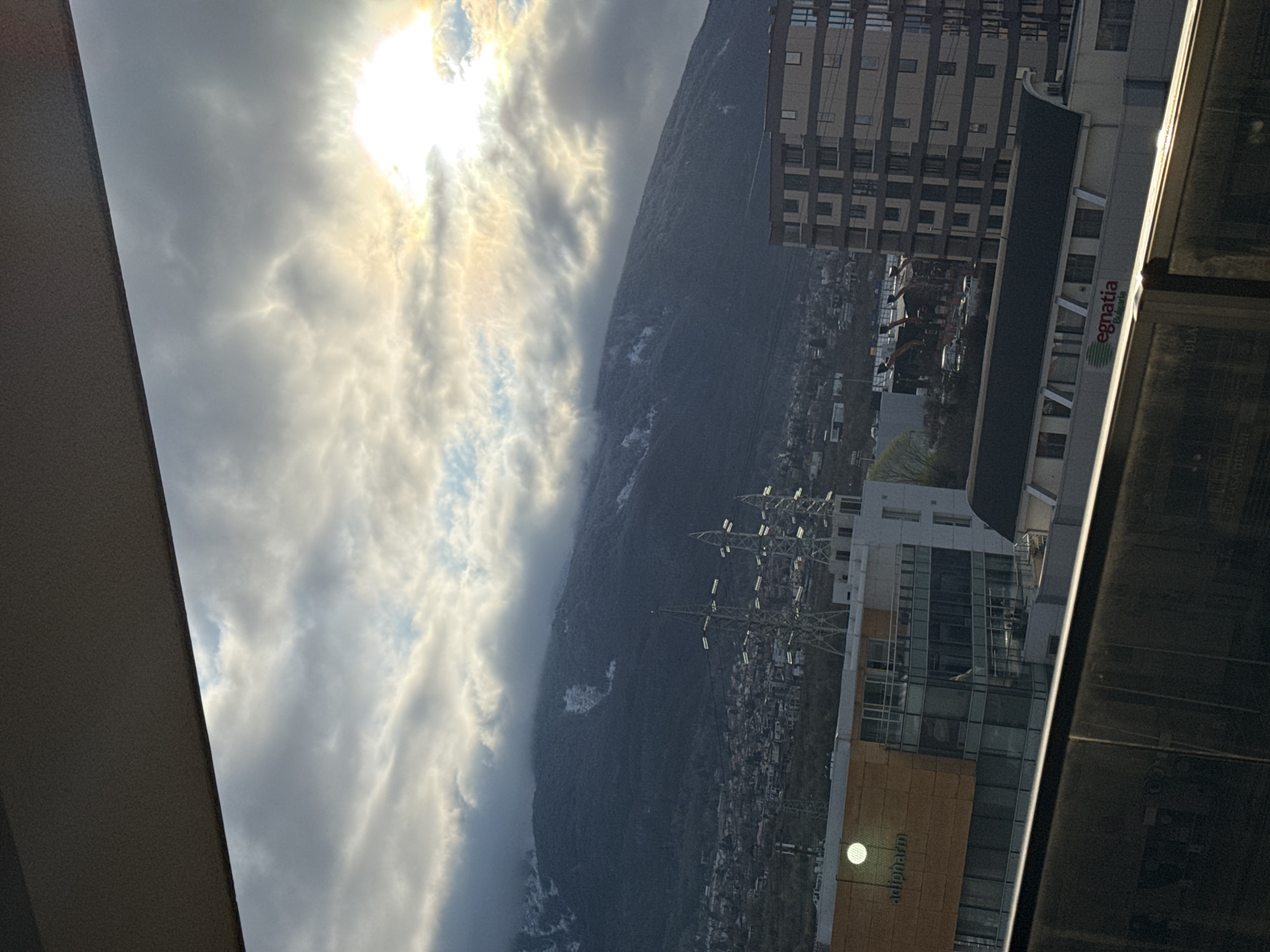 The image displays an urban landscape and mountainous backdrop, captured from an elevated vantage point, likely a balcony or window. The sky is partially cloudy, with scattered grey and white clouds. A bright sunburst is visible through the clouds in the upper right quadrant, suggesting either sunrise or sunset.
In the background, a large, dark mountain, identifiable as Vitosha Mountain, dominates the frame. Its upper slopes are partially covered in snow and intermittently obscured by low-hanging clouds.
The mid-ground reveals a sprawling urban area at the base of the mountain. Prominently featured are multiple lattice-structured electricity transmission towers (power pylons) carrying high-voltage lines. Below these, a light brown and white building with extensive glass windows is visible, bearing the "adipharm" logo on its facade along with a circular luminescent fixture. Further right and slightly lower, another building with a dark roof displays the "egnatia Bulgaria" logo. To the right, a modern, multi-story building with a patterned facade and numerous windows indicates recent construction or development. Various other residential and commercial structures, roads, and sparse vegetation are distributed throughout the urban sprawl.
The immediate foreground consists of a dark, reflective horizontal surface, consistent with a balcony railing or ledge, which partially obscures the bottom of the scene.
The location is Sofia, Bulgaria, with the view directed towards Vitosha Mountain, likely from an urban district such as Mladost, where companies like Adipharm are known to operate. No human figures are present. The scene conveys a cool, possibly early morning or late afternoon, atmosphere.