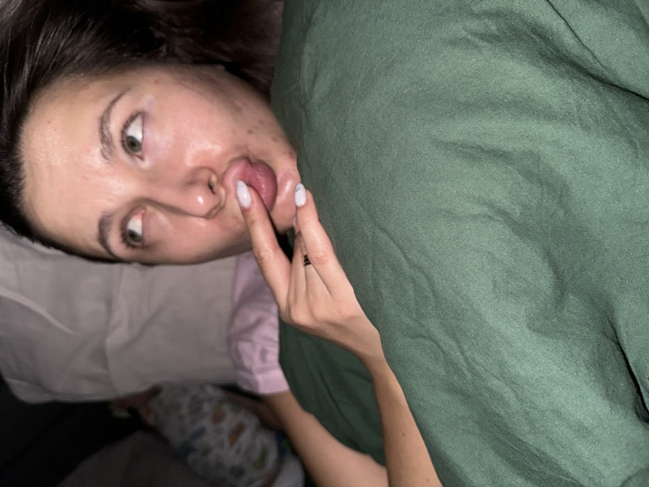 The image depicts an indoor scene in Molodi, Russia. A woman, appearing to be in her 20s or 30s, is positioned in the foreground, seemingly reclining on a bed. She has dark hair and light-colored eyes, with her gaze directed upwards and to the right. Her face exhibits a moist or oily appearance, and some blemishes are visible on her right cheek. Her lips are pursed, and her left index finger, displaying light-colored nail polish, is inserted between them, making contact with her visible tongue. A dark ring is present on her left ring finger. She is covered by a dark green fabric, likely a blanket or duvet, which shows creases. In the blurred background, to the left of the woman, a white pillow is partially visible, along with a small child or infant in patterned sleepwear and a section of pink fabric.