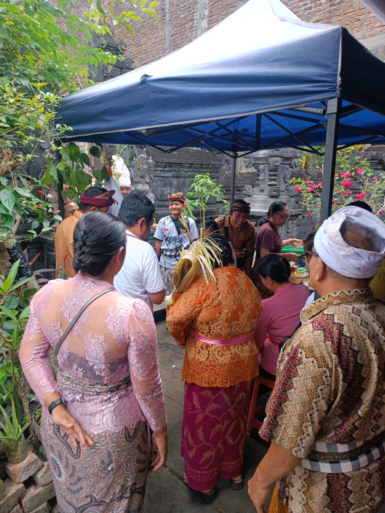 In Tabanan, Indonesia, a group of people are gathered outdoors under a blue and white canopy. The scene appears to be part of a cultural or religious event. Several individuals are wearing traditional Indonesian attire, including batik fabrics and sarongs. One person in the foreground, facing away from the camera, is dressed in a pink lace top and a patterned sarong, with a black watch on their wrist. Another person to their right wears a vibrant orange, intricately patterned outfit.
Behind them, men in white and brown attire are visible, some wearing headbands. One man in a white shirt is facing the group, while another, in a brown shirt and headband, smiles and gestures. A woman in a maroon top and glasses stands near him. Further in the background, stone carvings and lush green foliage suggest a temple or a traditional compound setting. Pink flowers bloom in the background. The ground is paved with stone. The overall atmosphere seems to be one of community and shared tradition.