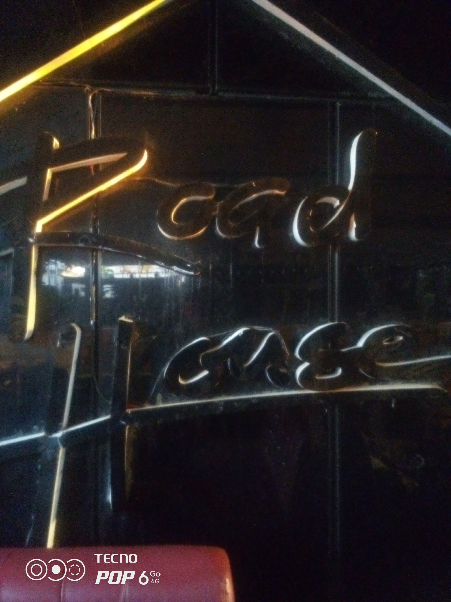 A black, reflective surface displays stylized, illuminated lettering, likely a sign. The text appears to read "Beach House," with the words stacked vertically. The letters are formed with a bright, glowing outline against the dark background. In the lower left corner, a partial view of a red seat is visible, along with a watermark from a TECNO POP 6 4G phone. The overall scene suggests an interior setting, possibly a bar, restaurant, or establishment with a distinct branding. The illumination and reflective material create a dramatic and modern aesthetic.