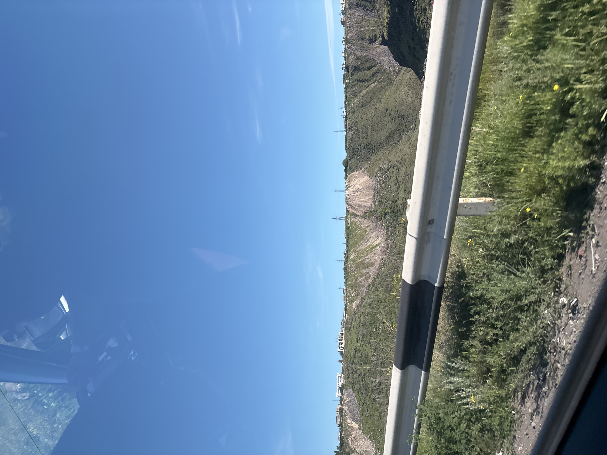The image depicts a view from inside a vehicle traveling along a road in what appears to be a mountainous or hilly area. The sky is a clear blue. On the right side of the frame is a guardrail, with black markings, beyond which there is green vegetation. Across from the guardrail, the landscape shows a slope with vegetation and what looks like houses at the top. It could be in the city of Kanakeravan, Armenia. The interior of the vehicle is visible on the lower left.
