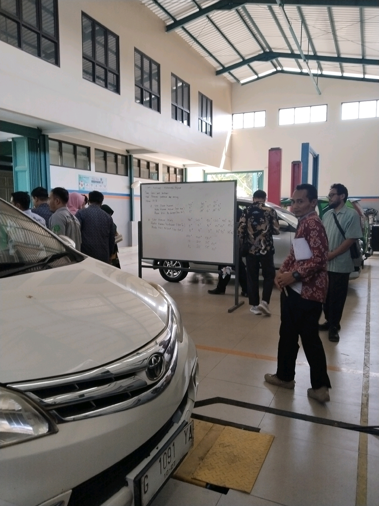 The image depicts an indoor automotive workshop environment, likely in Procol, Indonesia, given the license plate format and the presence of Indonesian writing on the whiteboard. A white car, possibly a Toyota Avanza based on the front grille and logo, is positioned in the foreground. Several individuals, appearing to be male, are present, some wearing traditional batik shirts and others casual attire. They are gathered around a whiteboard on wheels, which displays handwritten calculations and diagrams, suggesting an educational or diagnostic session. The workshop features a high ceiling with exposed industrial lighting and ventilation systems, common in such facilities. Windows are visible along the upper walls. The floor is tiled and marked with yellow and black lines.
